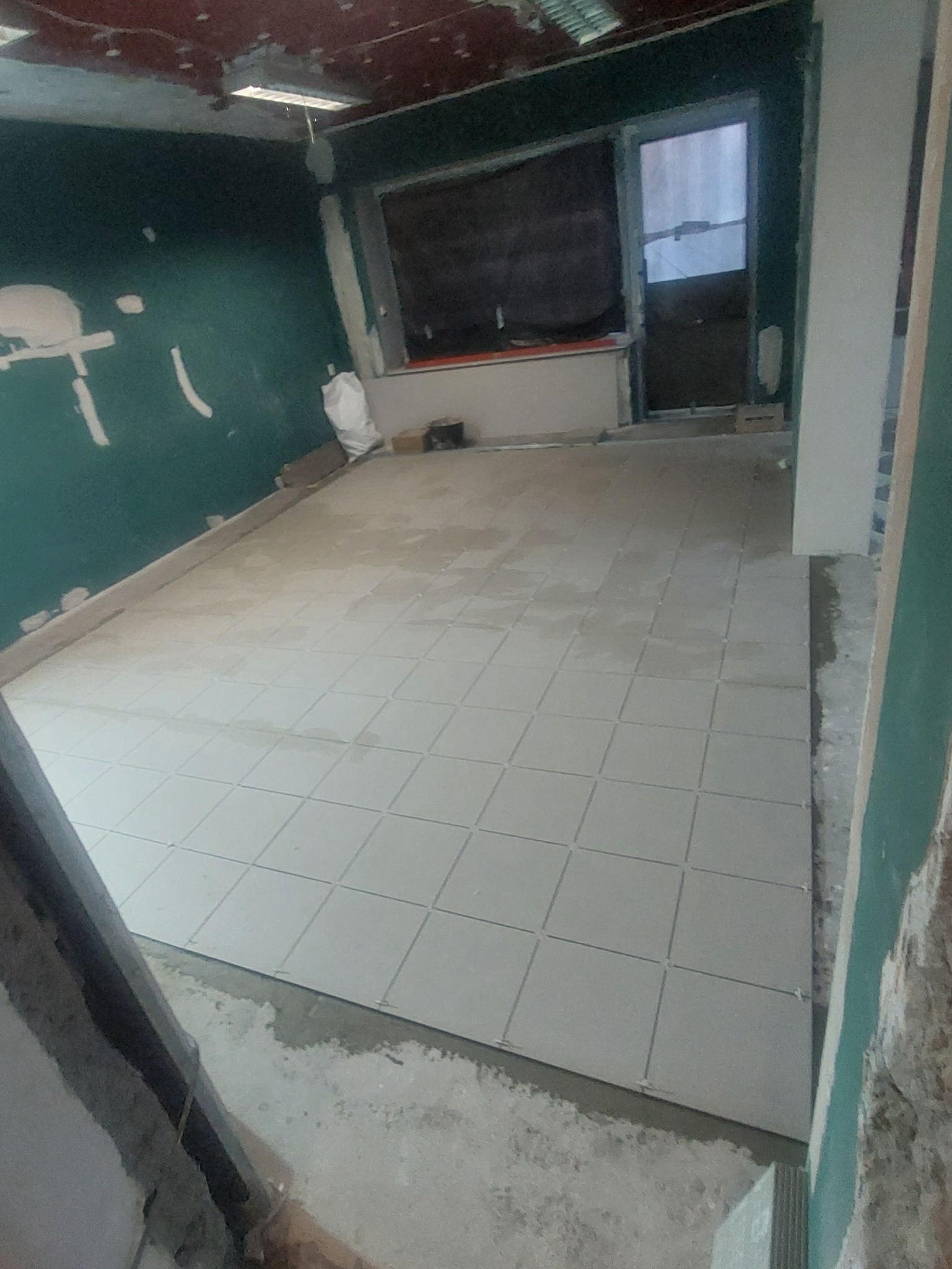 Wnętrze w trakcie remontu: ułożone jasne płytki podłogowe w kwadraty, ściany częściowo wykończone, widoczne niedoskonałości i worki z materiałami budowlanymi.