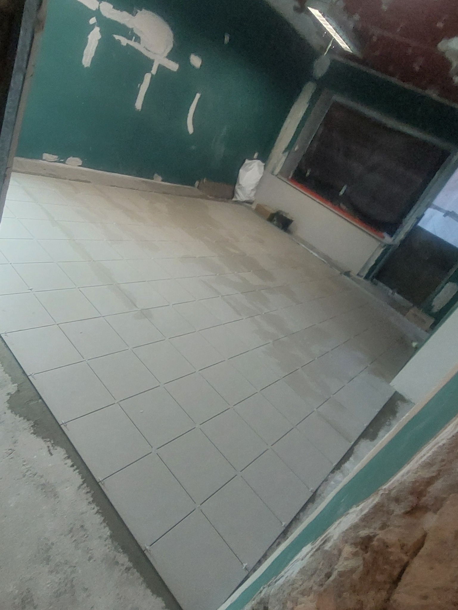 Układanie jasnych, kwadratowych płytek podłogowych w trakcie remontu. Ściana w kolorze butelkowej zieleni z widocznymi śladami szpachlowania. Nieukończone wykończenie.