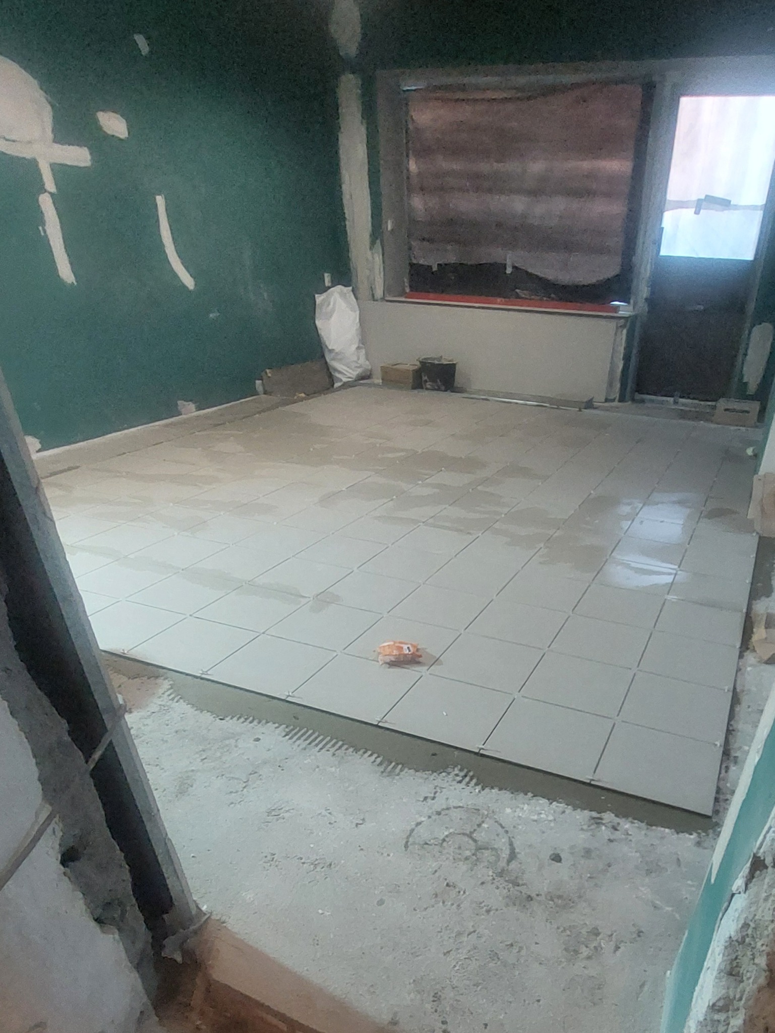 Wnętrze w trakcie remontu: ułożone jasne płytki podłogowe, ściany przygotowane do malowania, widoczny worek z zaprawą. Fragment widoku na okno.