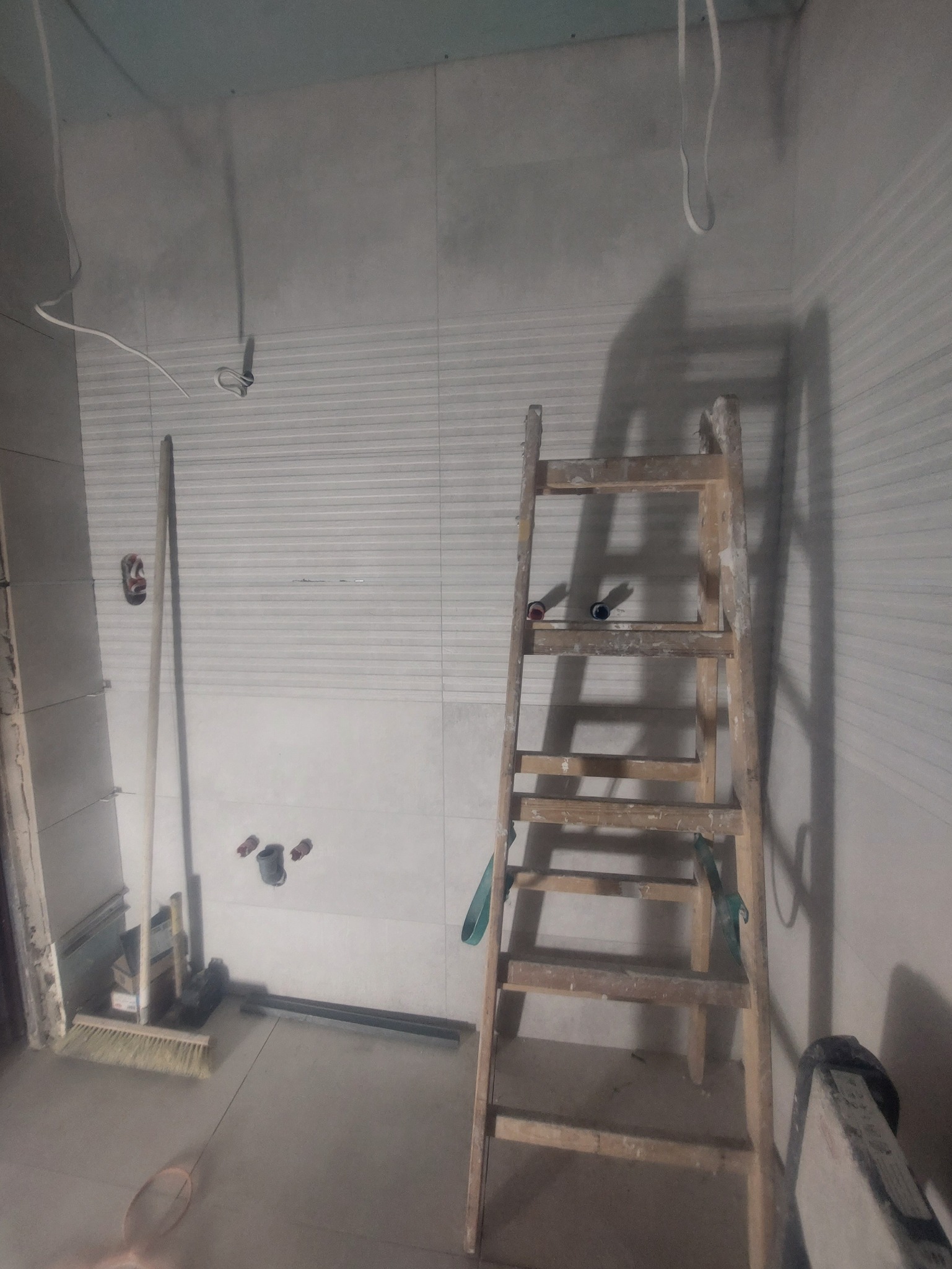 Wnętrze łazienki w trakcie prac wykończeniowych. Widoczna drabina oparta o ścianę z częściowo ułożonymi płytkami, instalacja wodna i narzędzia. W tle sufit z płyt karton-gips.
