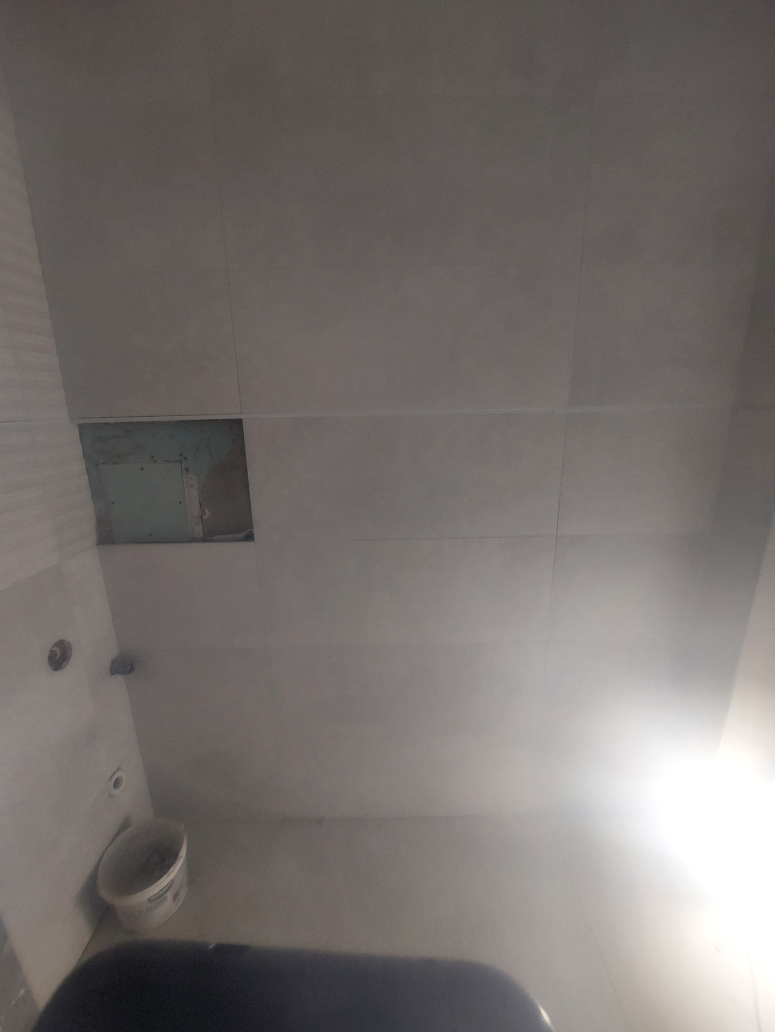 Ściana w łazience z jasnymi płytkami, widoczne niedokończone miejsce po płytce i wiadro. Oświetlenie od dołu, perspektywa z dołu do góry.