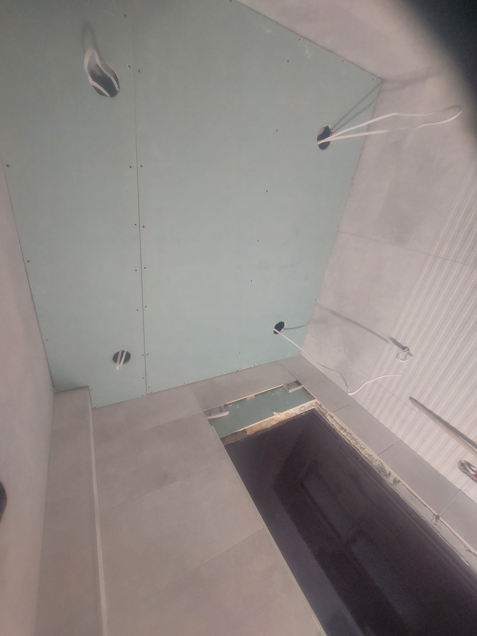 Montaż sufitu z płyty GK w łazience, widoczne otwory na oświetlenie i wystające kable. Płytki na ścianach, przygotowanie do wykończenia.