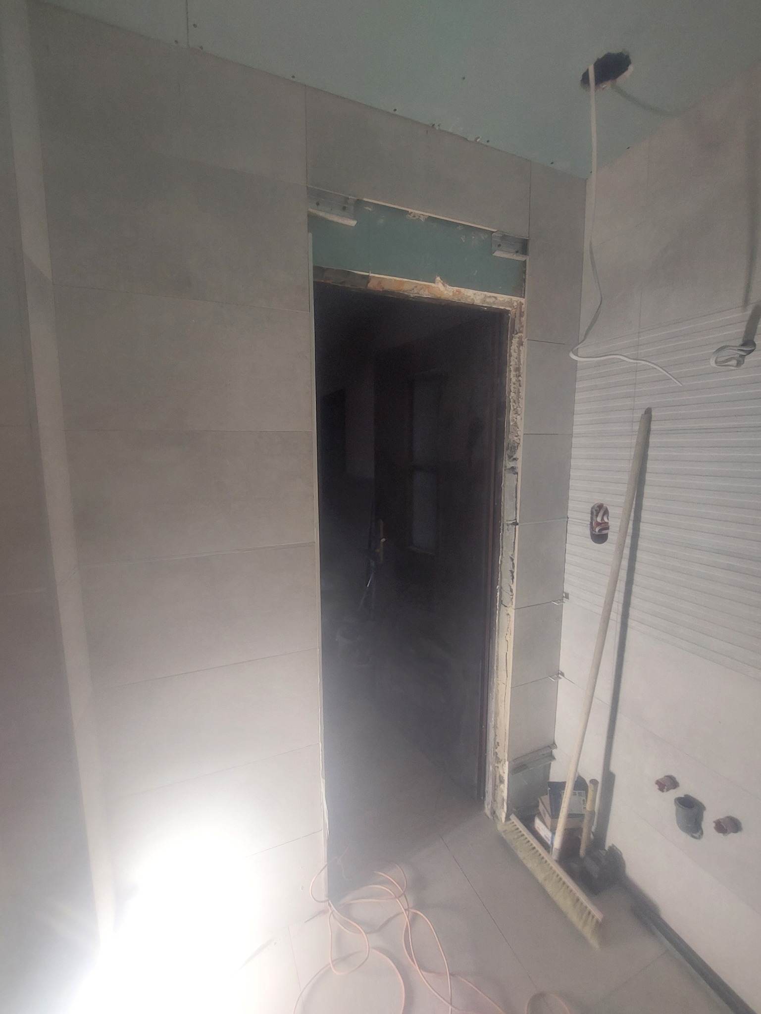 Wnętrze łazienki w trakcie remontu: widoczne płytki na ścianach, przygotowane do montażu drzwi, przewody i narzędzia budowlane. Etap prac wykończeniowych.