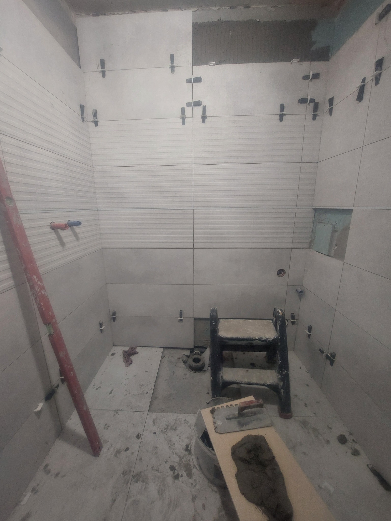 Montaż płytek w łazience z użyciem krzyżyków dystansowych. Widoczna drabinka, poziomica i narzędzia glazurnicze. Częściowo wykończone ściany z poziomymi wzorami.