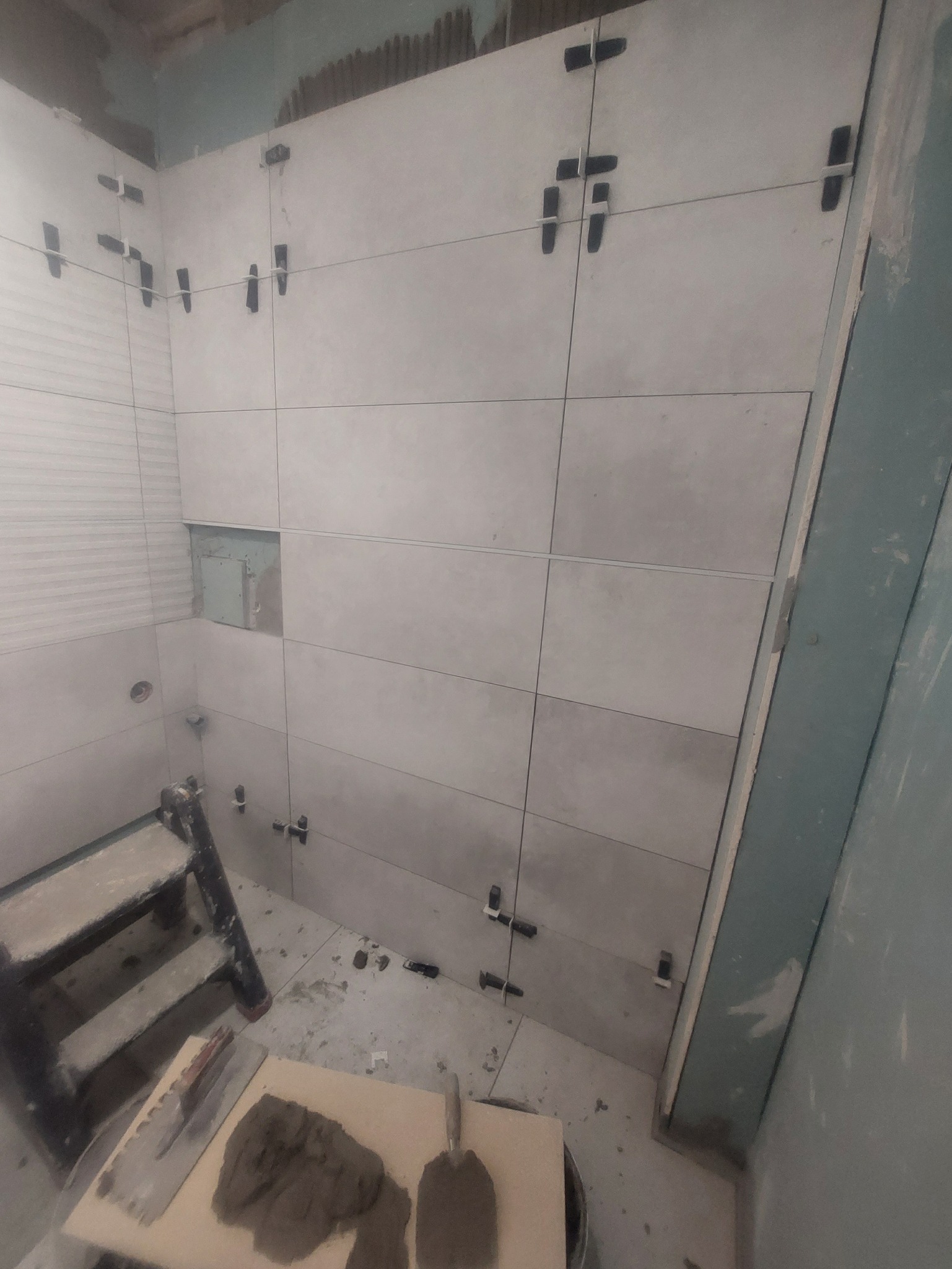 Ściana łazienki w trakcie wykańczania z szarymi płytkami, narzędziami i drabinką, widoczne klipsy poziomujące płytki.