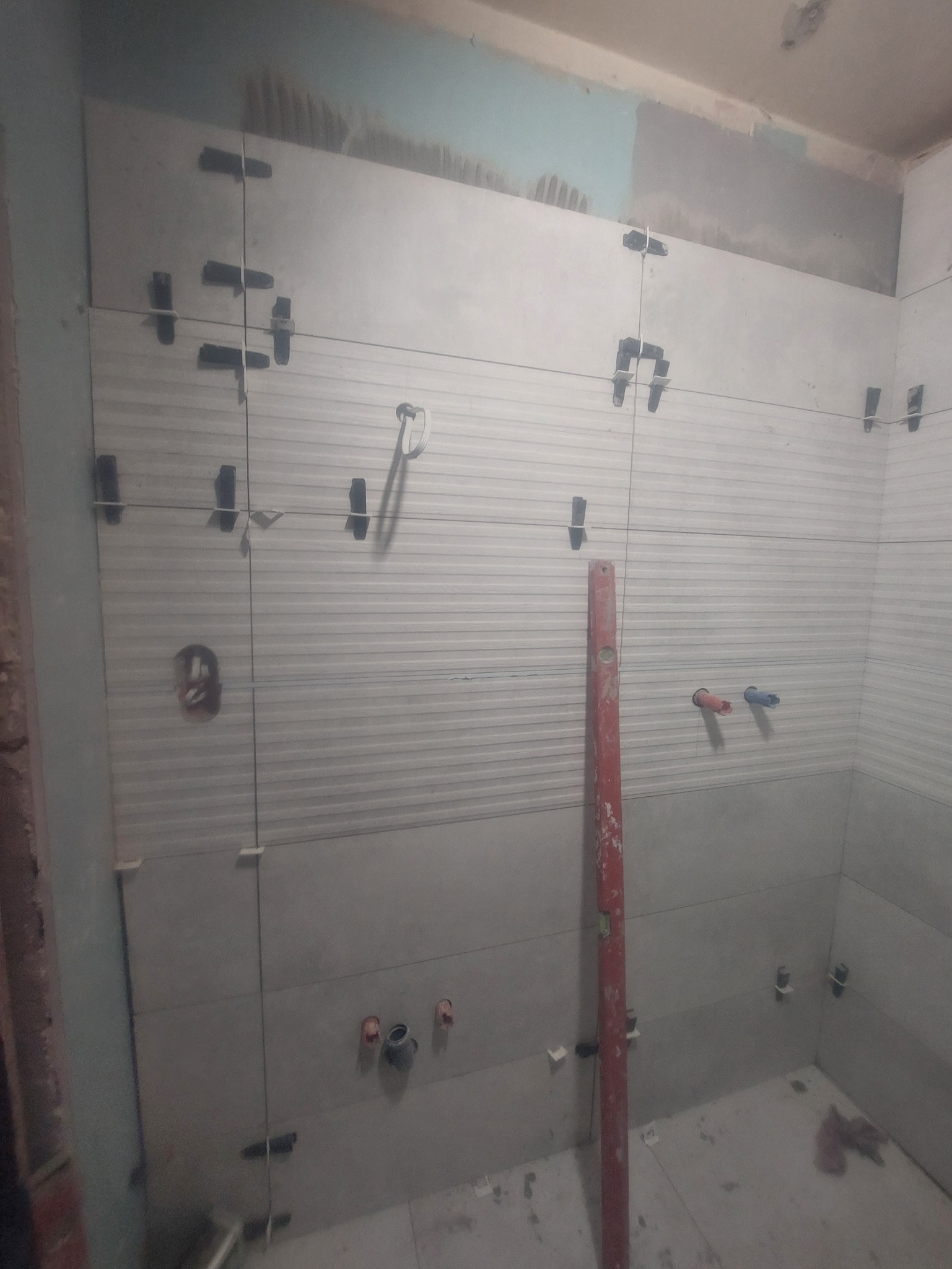 Układanie jasnych, poziomych płytek z fakturą w łazience. Widoczne krzyżyki dystansowe, poziomica i fragment instalacji. W tle surowy fragment ściany.