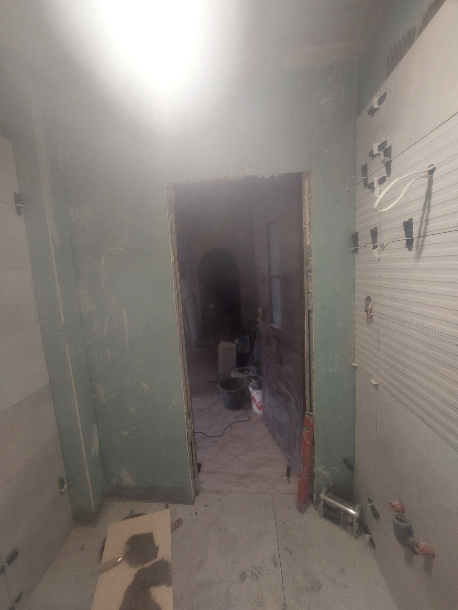 Remont łazienki: świeżo położone płytki na ścianie, widoczne narzędzia i materiały budowlane, surowa ściana w tle. Prace w toku, widok na sąsiednie pomieszczenie.