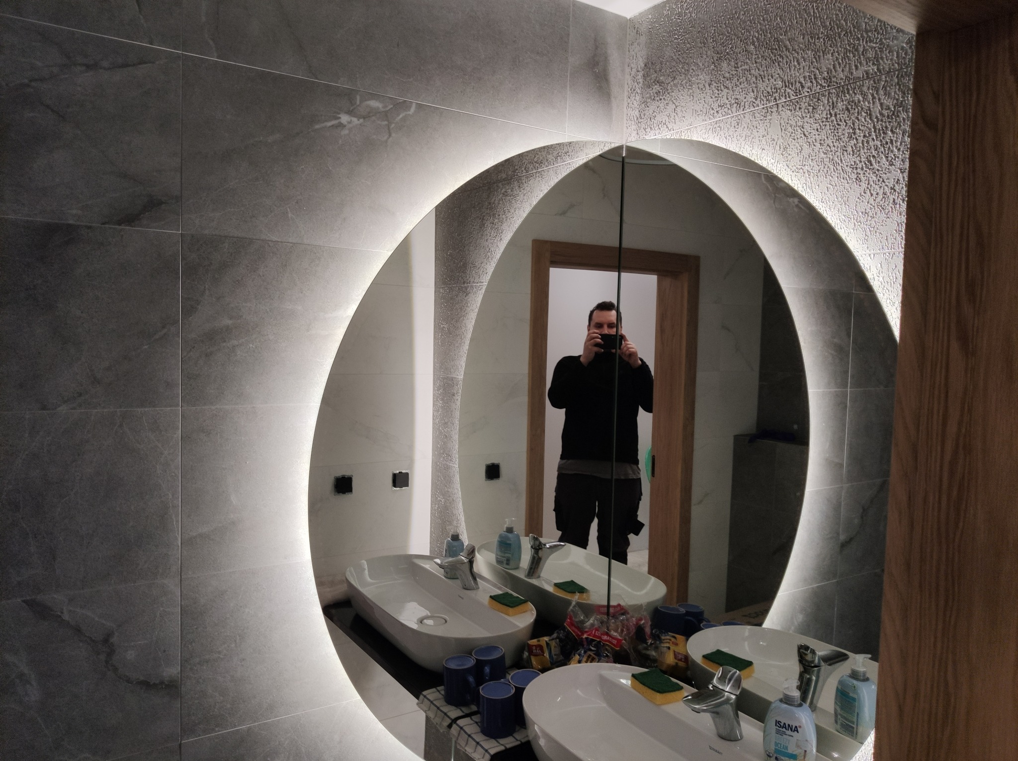 Nowoczesna łazienka z podświetlanym lustrem w kształcie elipsy, szare płytki imitujące kamień na ścianach, dwie białe umywalki nablatowe, widoczne puszki elektryczne bez osprzętu na ścianie...