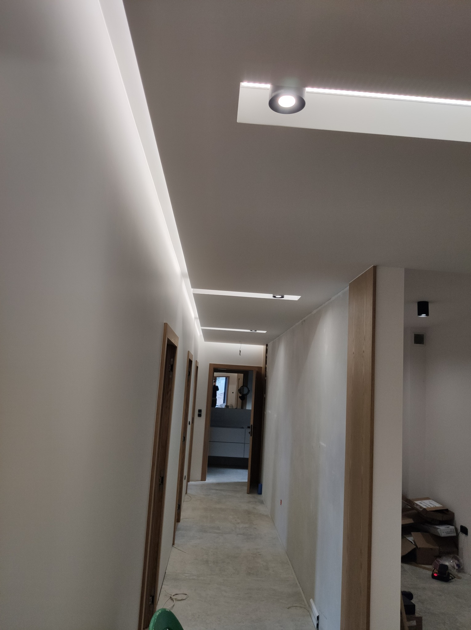 Korytarz w trakcie wykańczania z zamontowanym oświetleniem LED w suficie podwieszanym i punktowymi lampami sufitowymi, widoczne drzwi wewnętrzne i kartony w tle.