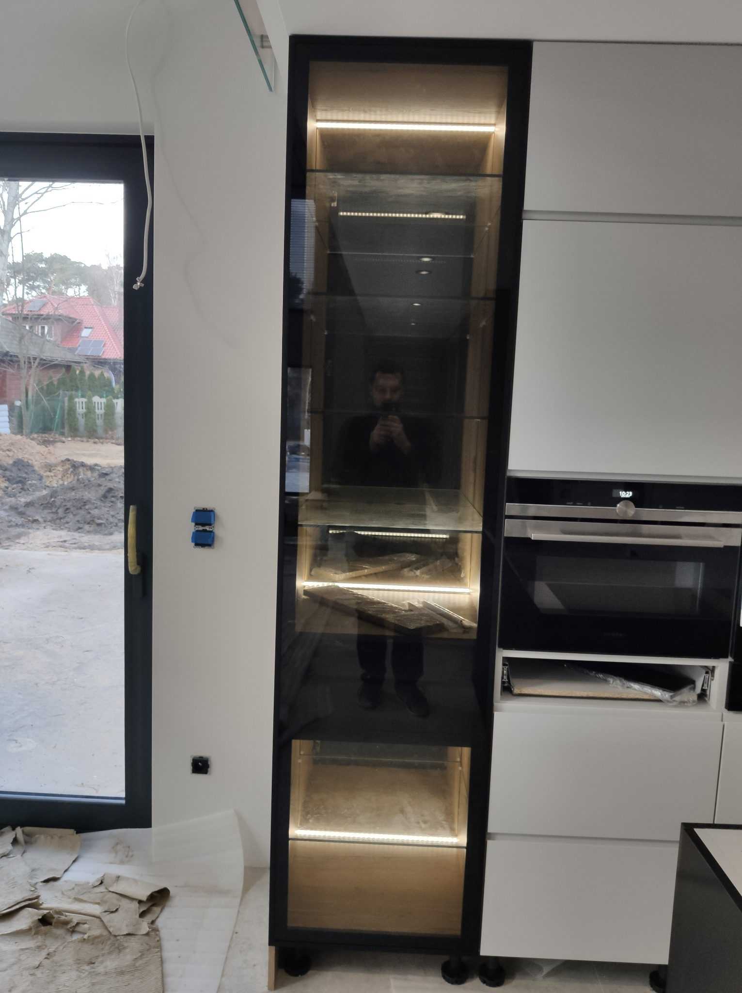 Montaż oświetlenia LED w szklanej witrynie kuchennej, widoczne podłączenie elektryczne i odbicie instalatora w szybie.