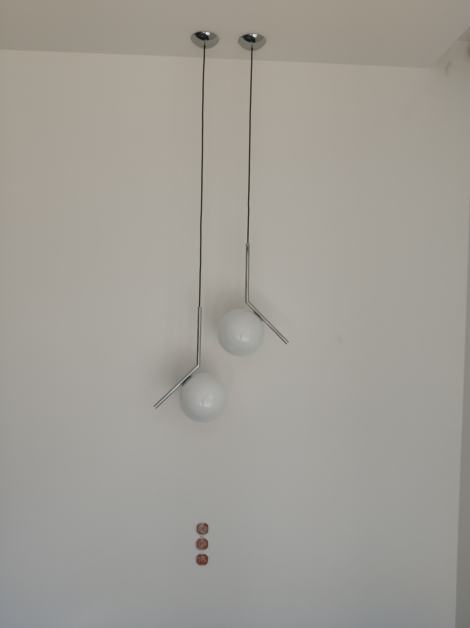 Dwa nowoczesne, białe, kuliste klosze lamp wiszących na cienkich, czarnych przewodach, zamocowane do sufitu za pomocą chromowanych podsufitek. Na ścianie widoczne ślady po otworach montażowych.