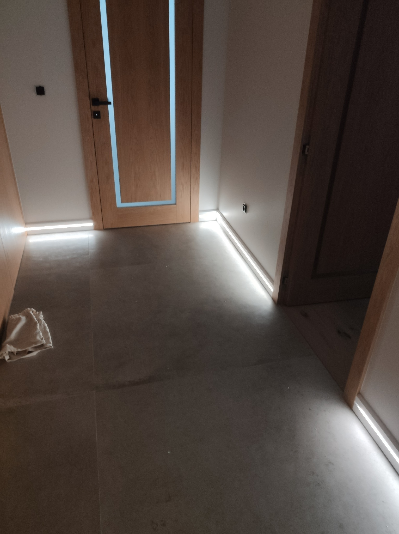 Korytarz we Włocławku z zamontowanym oświetleniem LED przypodłogowym, widoczne dwie drewniane ościeżnice drzwiowe z czarnymi klamkami oraz szara podłoga z gresu.