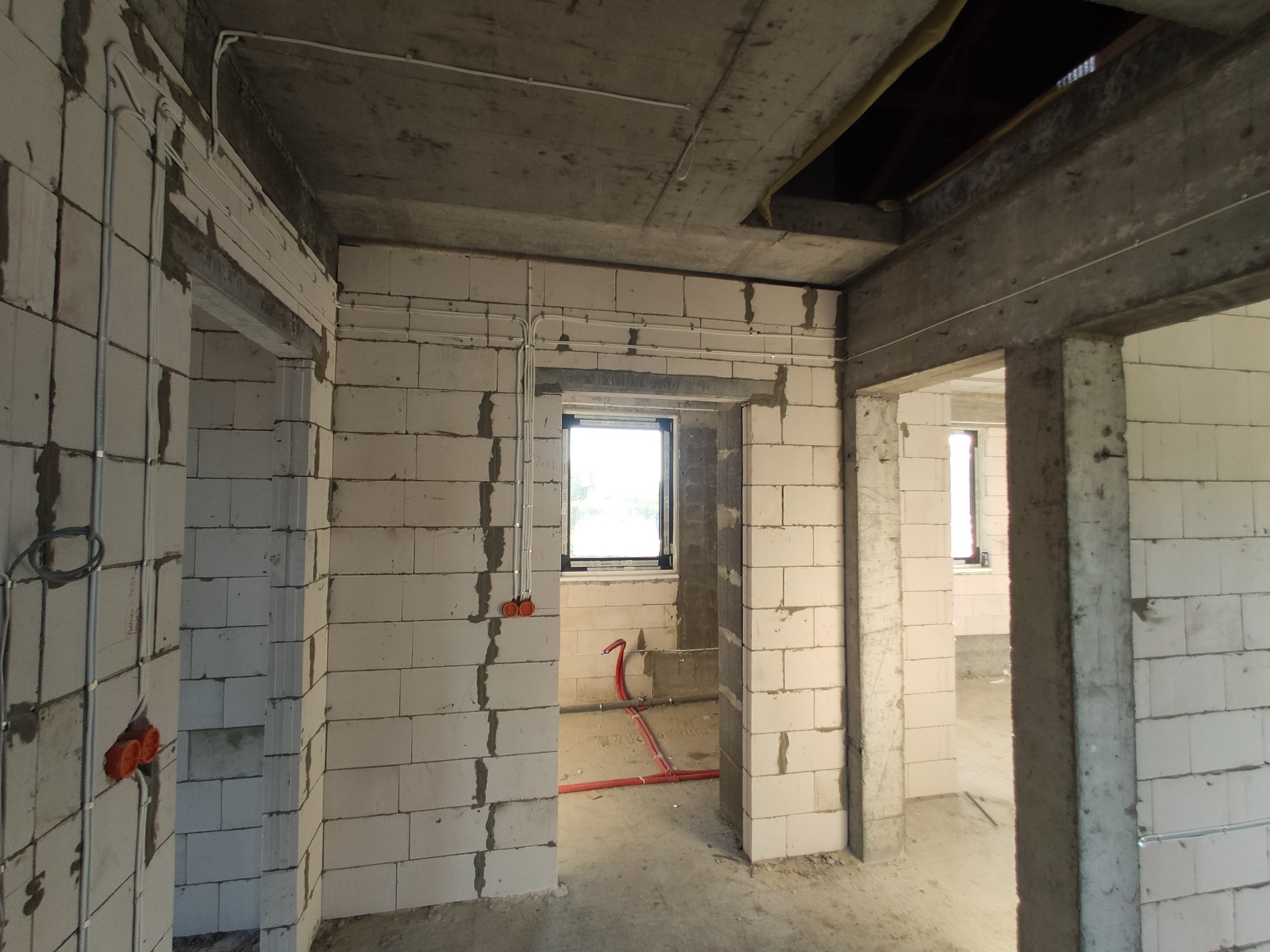 Surowy stan wnętrza budynku w trakcie prac instalacyjnych: widoczne białe bloczki, betonowy strop, rozprowadzone kable elektryczne w peszlach i puszkach, instalacja wodna w czerwonych rurach.