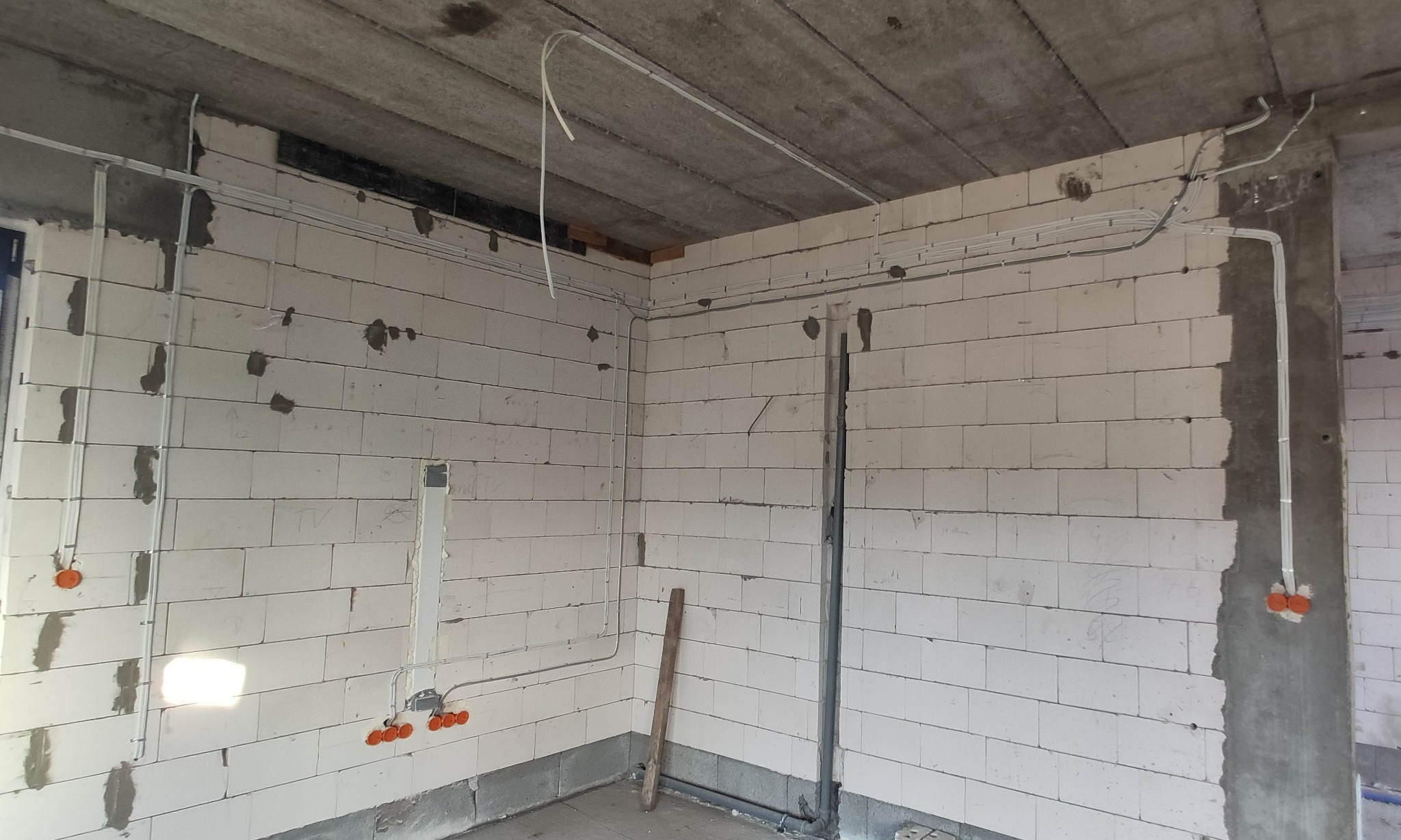 Surowy stan wnętrza z białymi bloczkami, widoczne szare rury instalacyjne i pomarańczowe puszki elektryczne, kable rozprowadzone po ścianach i suficie.