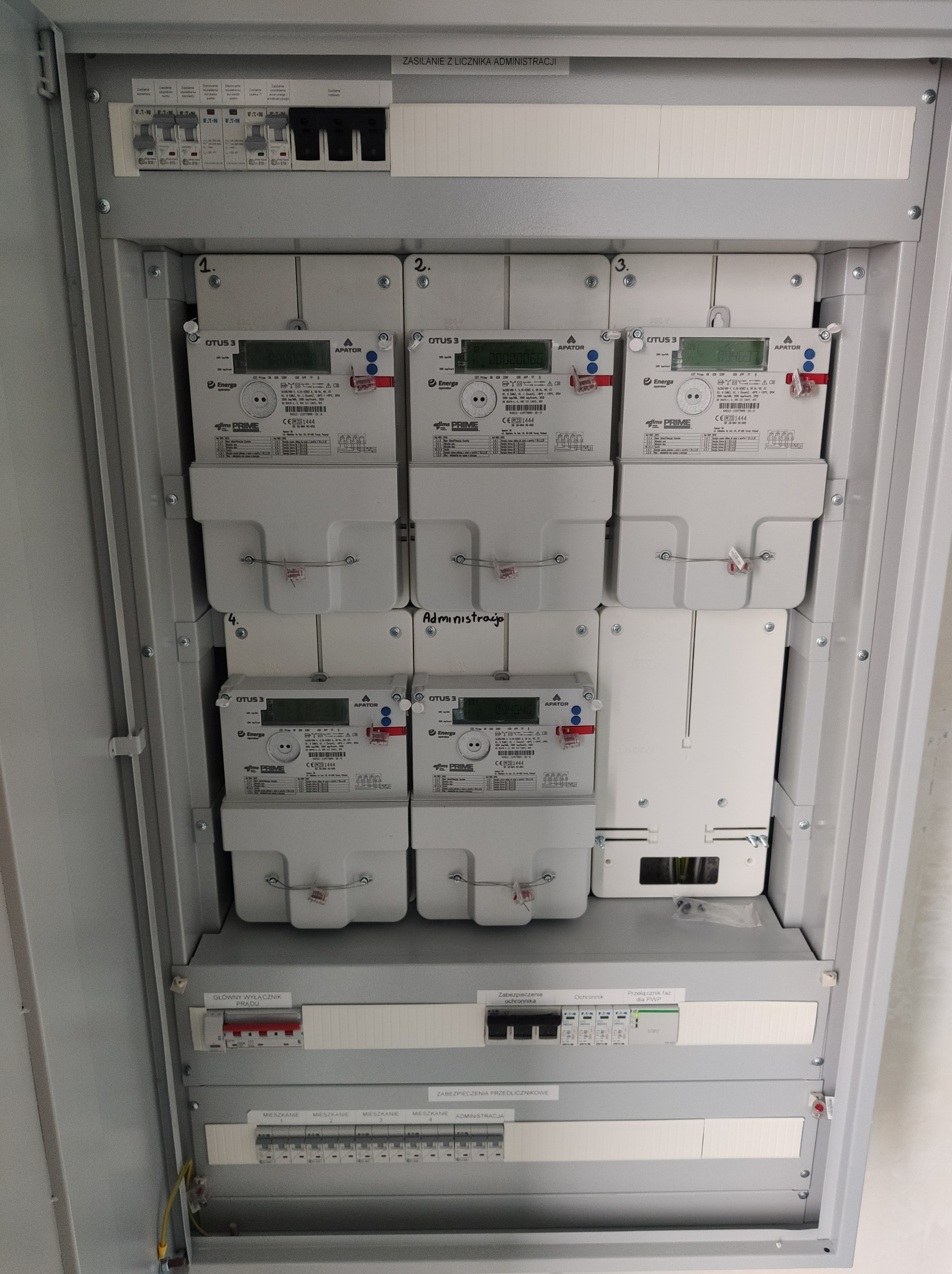Rozdzielnica elektryczna z zamontowanymi licznikami energii elektrycznej, widoczne zabezpieczenia nadprądowe i wyłączniki różnicowoprądowe, etykiety opisujące obwody.