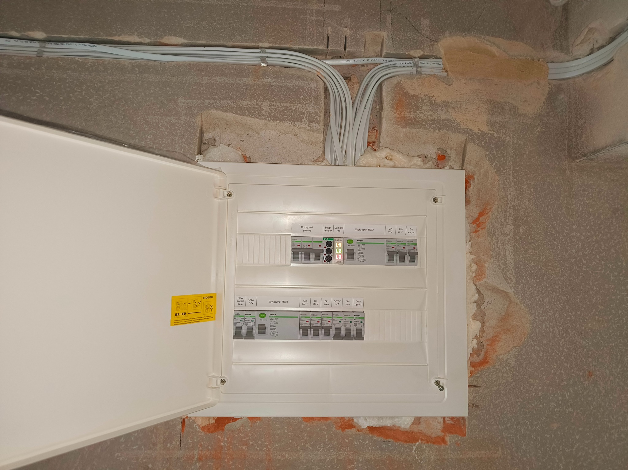 Otwarta skrzynka bezpiecznikowa firmy Noark zamontowana na surowej ścianie, widoczne białe kable elektryczne prowadzone po suficie.