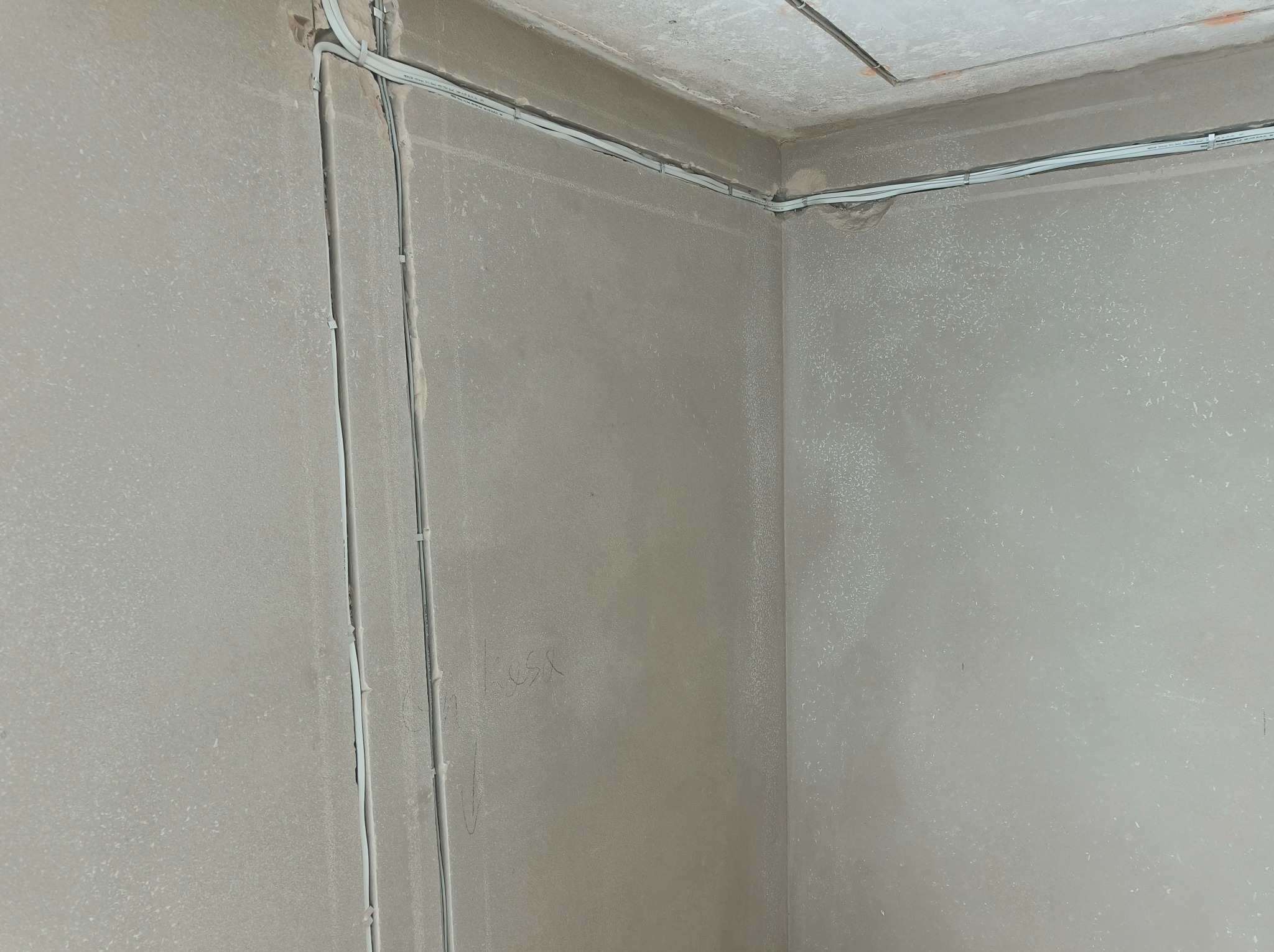 Surowe wnętrze w trakcie prac wykończeniowych, widoczne kable elektryczne prowadzone po ścianach i pod sufitem, przytwierdzone uchwytami, ściany w szarym tynku.