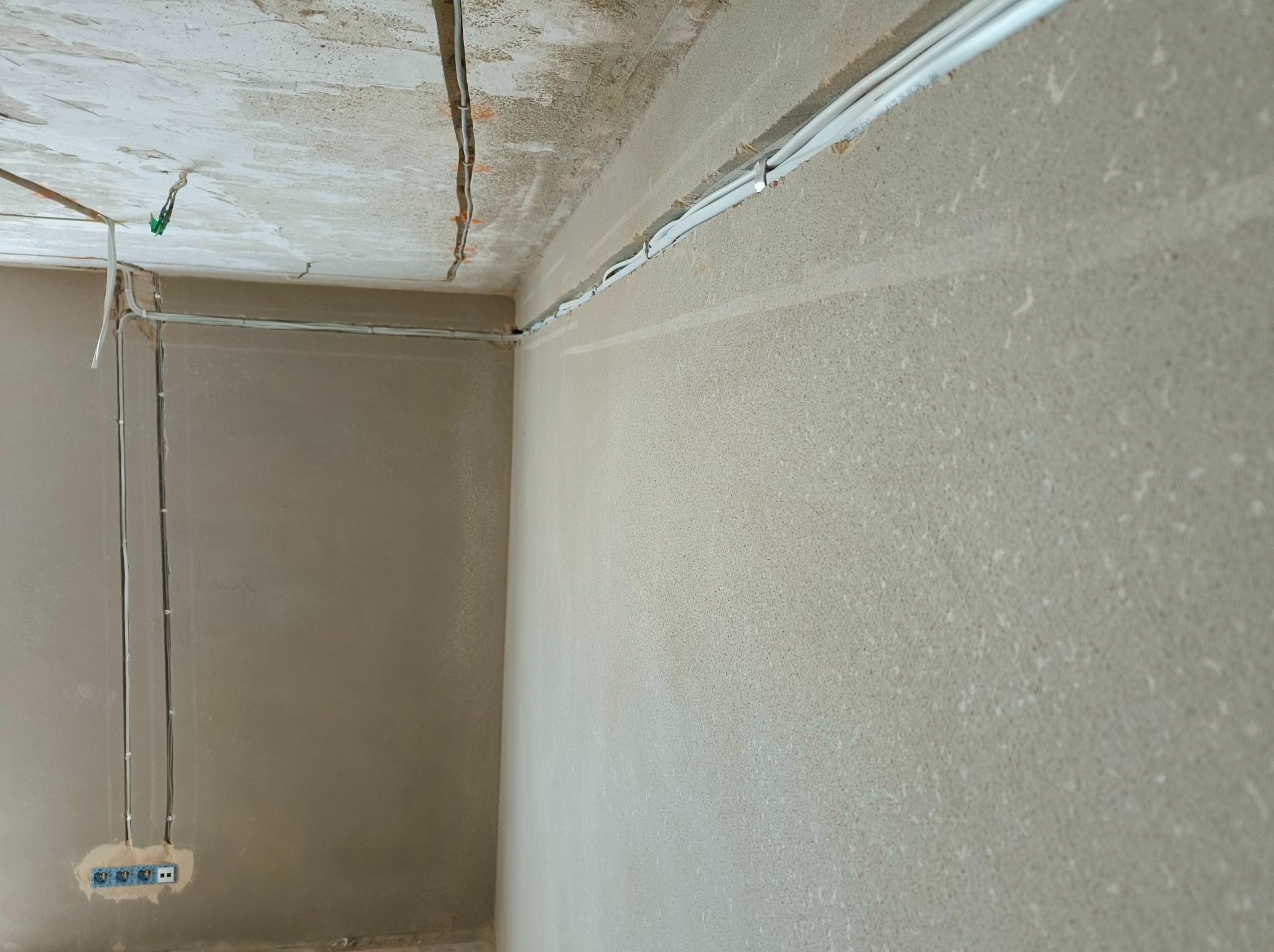 Szare wnętrze w trakcie remontu z widocznymi przewodami elektrycznymi prowadzonymi po ścianach i suficie, mocowane uchwytami, z zamontowanym gniazdkiem elektrycznym.