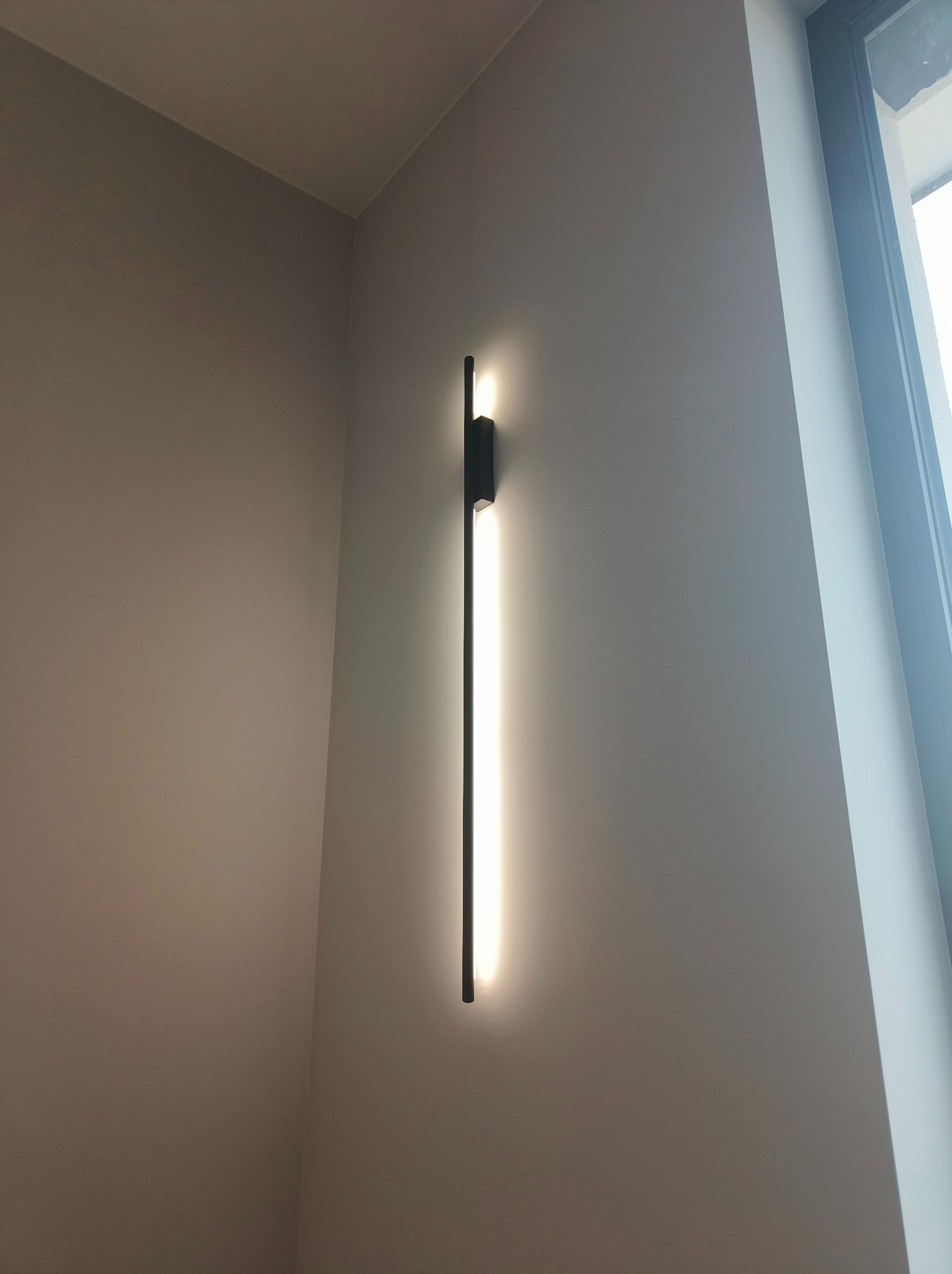 Minimalistyczna, pionowa lampa ścienna LED z czarną obudową, oświetlająca szarą ścianę w pobliżu okna z widocznym fragmentem ramy.