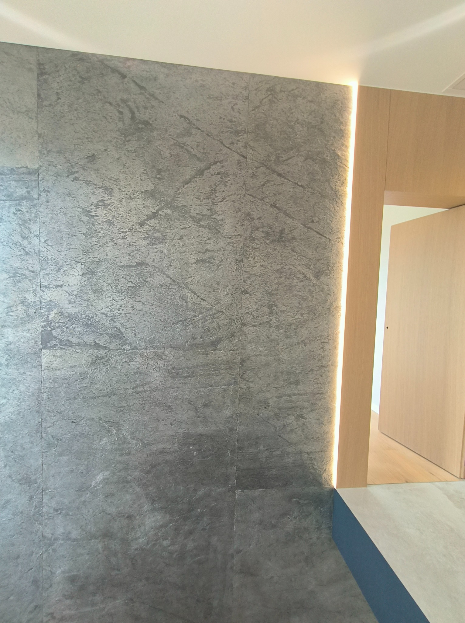 Ściana wykończona szarymi płytami o teksturze kamienia z wbudowanym pionowym oświetleniem LED, sąsiadująca z drewnianą framugą drzwi prowadzących do jasnego pomieszczenia.