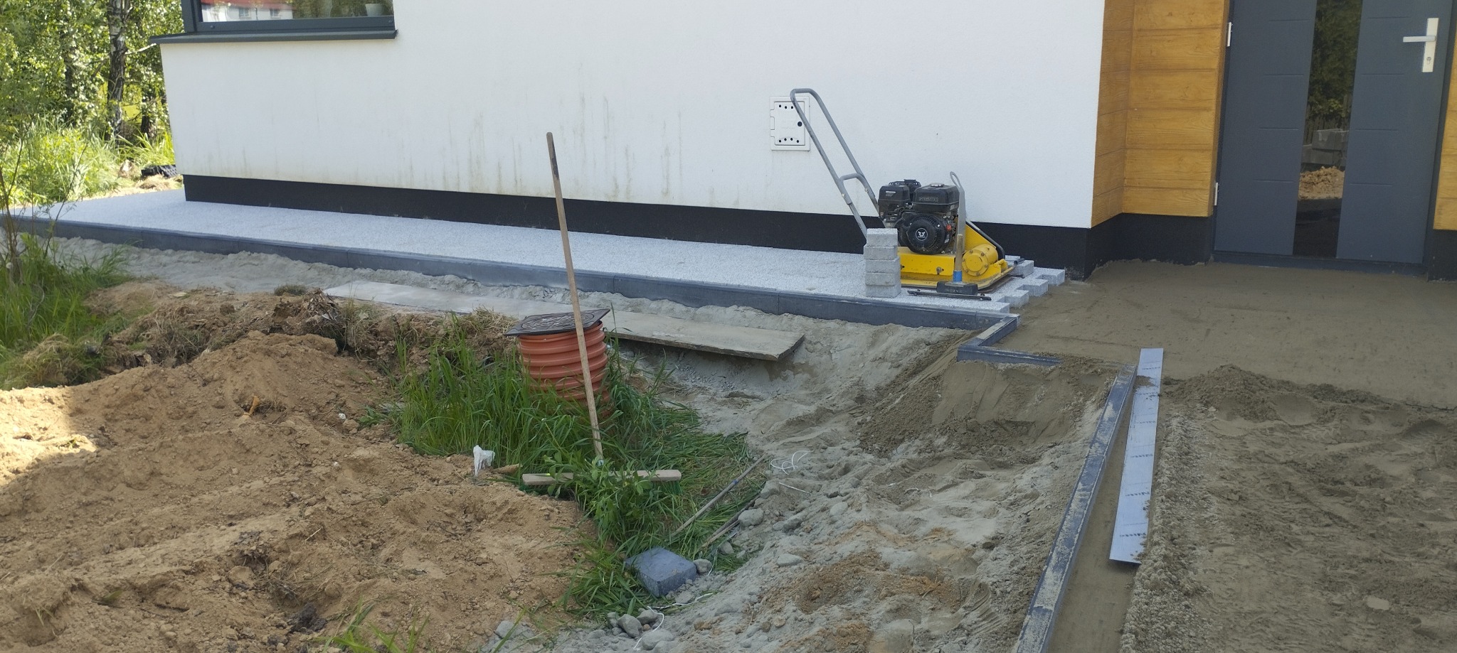 Układanie kostki brukowej przy domu: ubijarka spalinowa na warstwie podsypki obok ułożonych obrzeży. Widoczny stos kostki brukowej i wykop pod przyszły chodnik.
