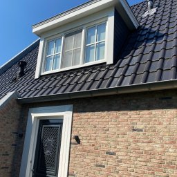 White Crow Usługi Remontowo-Budowlane Jacek Gdaniec - Ceglana elewacja domu z czarną dachówką, białymi obramowaniami okien i drzwi oraz metalowym kominem na dachu w słoneczny dzień. Widok z dołu.