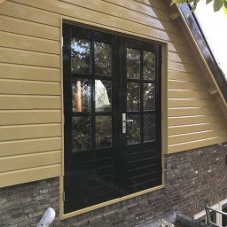 White Crow Usługi Remontowo-Budowlane Jacek Gdaniec - Odnowiona elewacja budynku z drewnianą okładziną w odcieniach beżu i czarnymi dwuskrzydłowymi drzwiami, widoczne narzędzia malarskie i ceglana podmurówka.