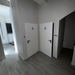 White Crow Usługi Remontowo-Budowlane Jacek Gdaniec - Wnętrze po malowaniu: białe ściany, szara podłoga, dwie otwarte toalety z czarnymi klamkami, widoczny fragment okna i słupka do tańca.