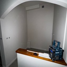 White Crow Usługi Remontowo-Budowlane Jacek Gdaniec - Wnęka po malowaniu na biało z radiem Makita na drewnianym blacie. Widoczny fragment instalacji elektrycznej i wentylacji. Świeżo pomalowane ściany z łukowym przejściem.