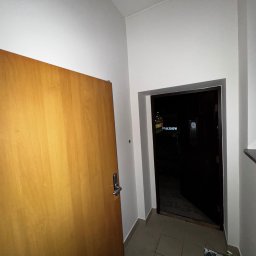 White Crow Usługi Remontowo-Budowlane Jacek Gdaniec - Wnętrze klatki schodowej po odświeżeniu: białe ściany, drewniane drzwi wejściowe i widok na zewnątrz przez otwarte drzwi. Na podłodze płytki i wiadro z narzędziami.