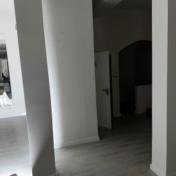 White Crow Usługi Remontowo-Budowlane Jacek Gdaniec - Białe, świeżo pomalowane ściany i kolumny w jasnym wnętrzu. Na podłodze panele imitujące drewno. W głębi widoczne drzwi. W rogu leży lampa budowlana.
