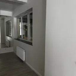 Wnętrze po odświeżeniu: gładkie, białe ściany, okno z parapetem, grzejnik. Podłoga z paneli. Widok na sąsiednie budynki odbija się w oknie i lustrze.