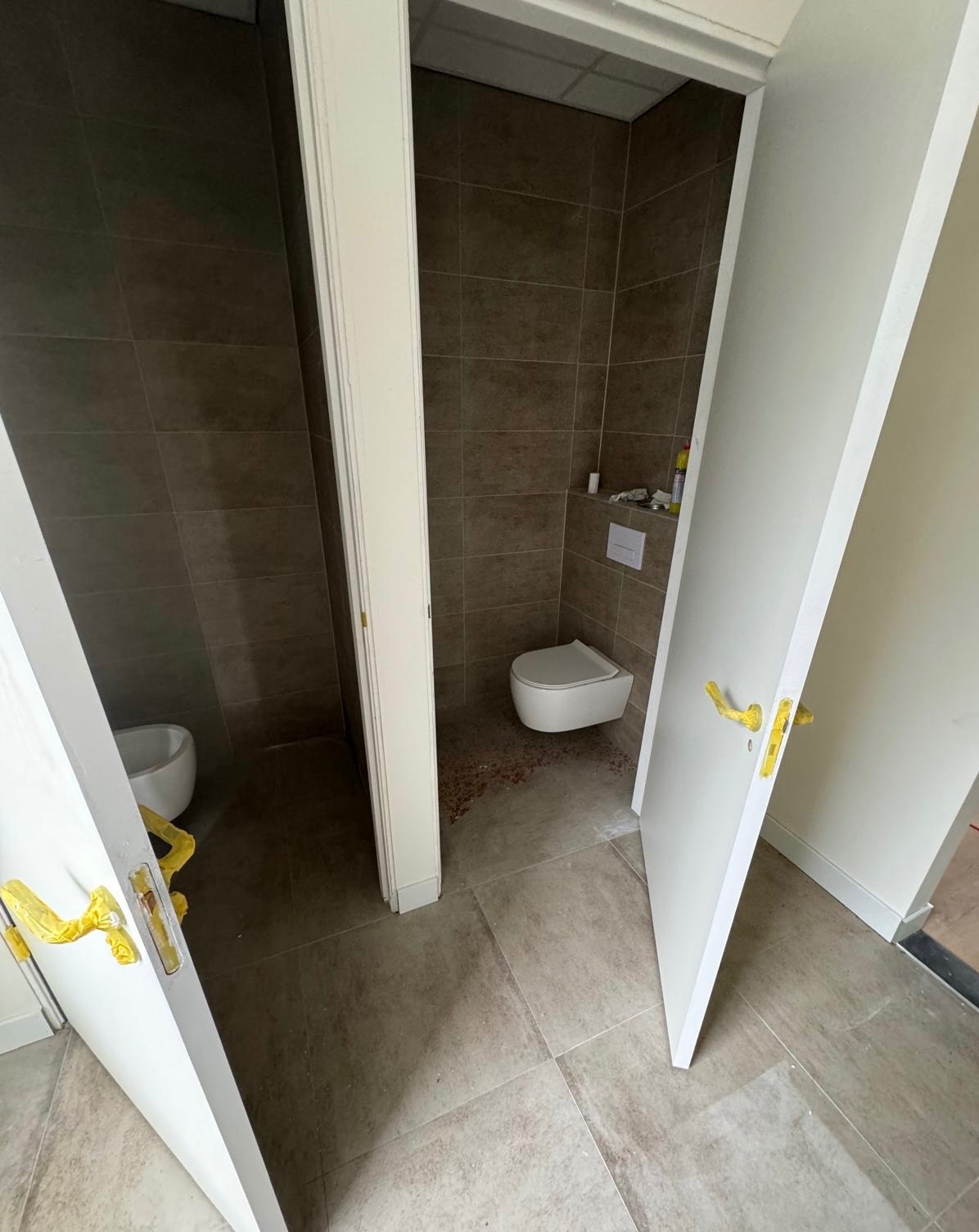 Dwa otwarte, białe drzwi prowadzą do wykafelkowanych łazienek. Na podłodze płytki, widoczne elementy instalacji. Drzwi zabezpieczone żółtą taśmą.