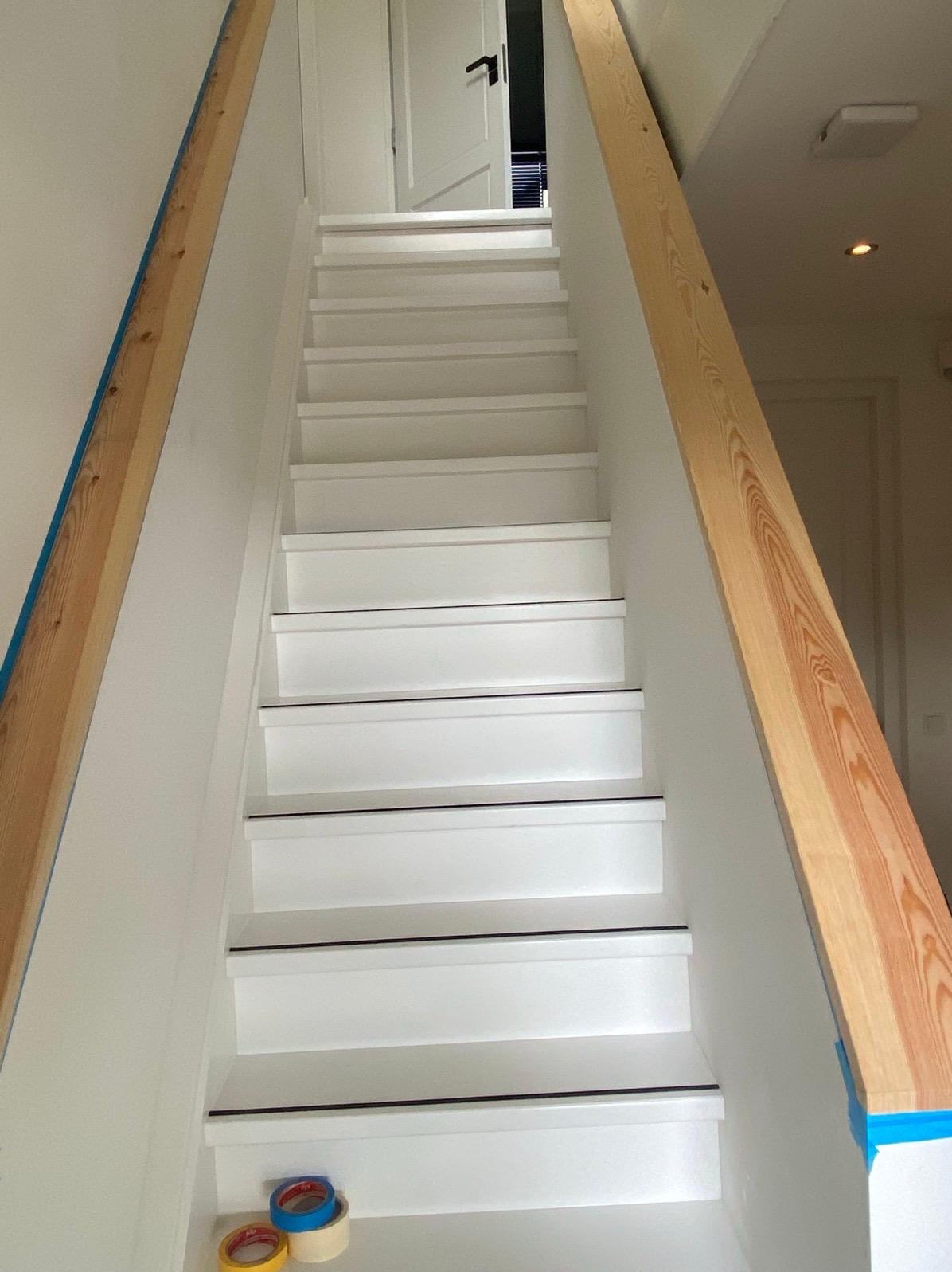 Białe schody z drewnianymi poręczami zabezpieczonymi taśmą malarską, widok z dołu. Taśmy malarskie leżące na dole schodów. Remont klatki schodowej.
