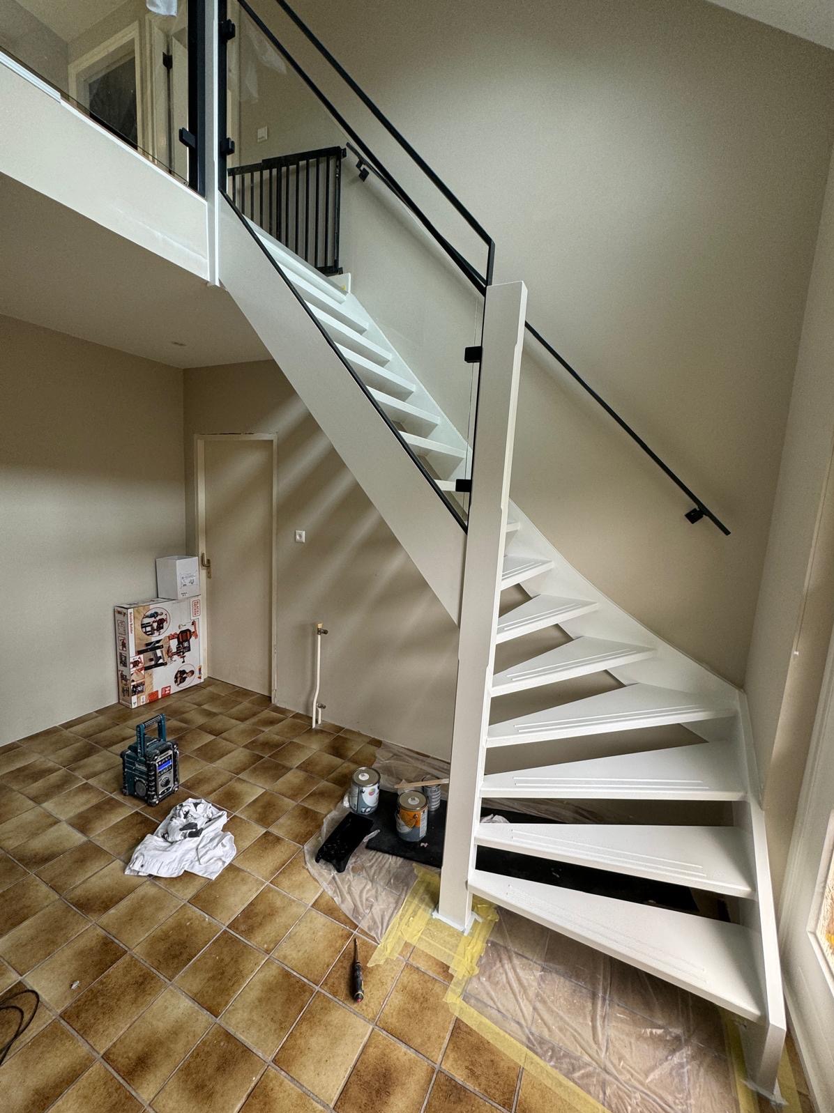 Białe schody z czarną balustradą po malowaniu w jasnym wnętrzu. Widoczne puszki z farbą, narzędzia i zabezpieczona podłoga. Ujęcie z dołu, podkreśla geometrię schodów.