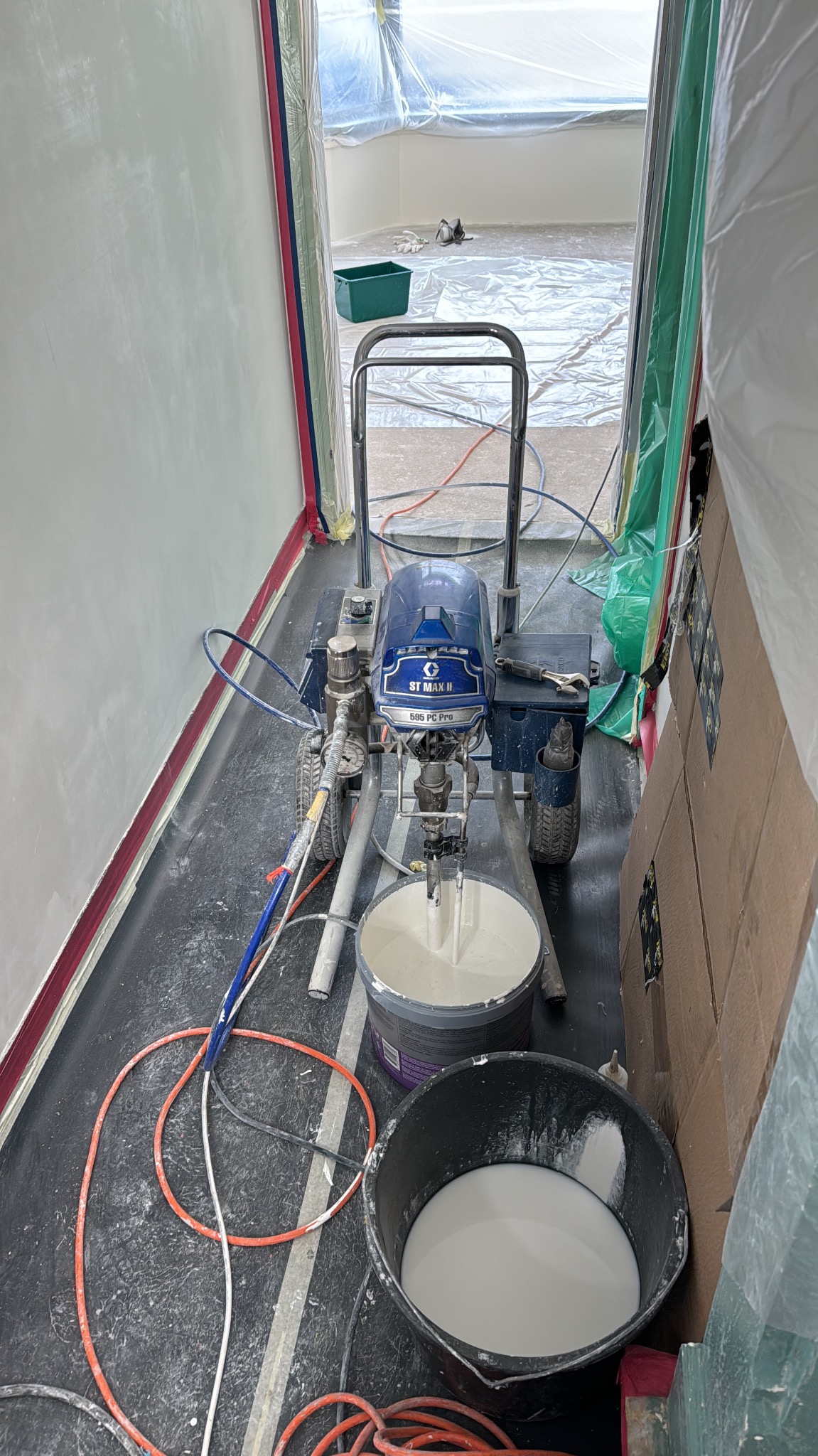 Agregat malarski ST MAX II 595 PC Pro z wiaderkami farby w pomieszczeniu zabezpieczonym folią malarską, gotowy do natryskowego malowania ścian.