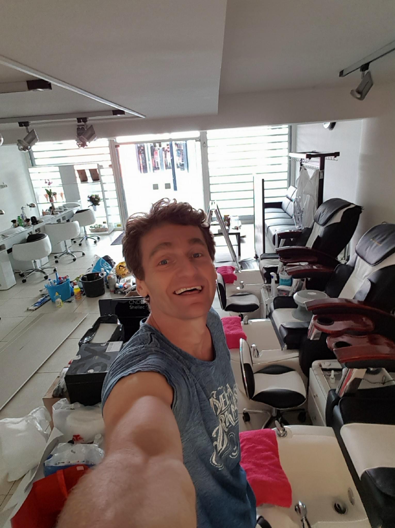 Mężczyzna robi selfie w salonie kosmetycznym przed remontem. Widoczne stanowiska do manicure, fotele do pedicure i bałagan po pracach przygotowawczych.