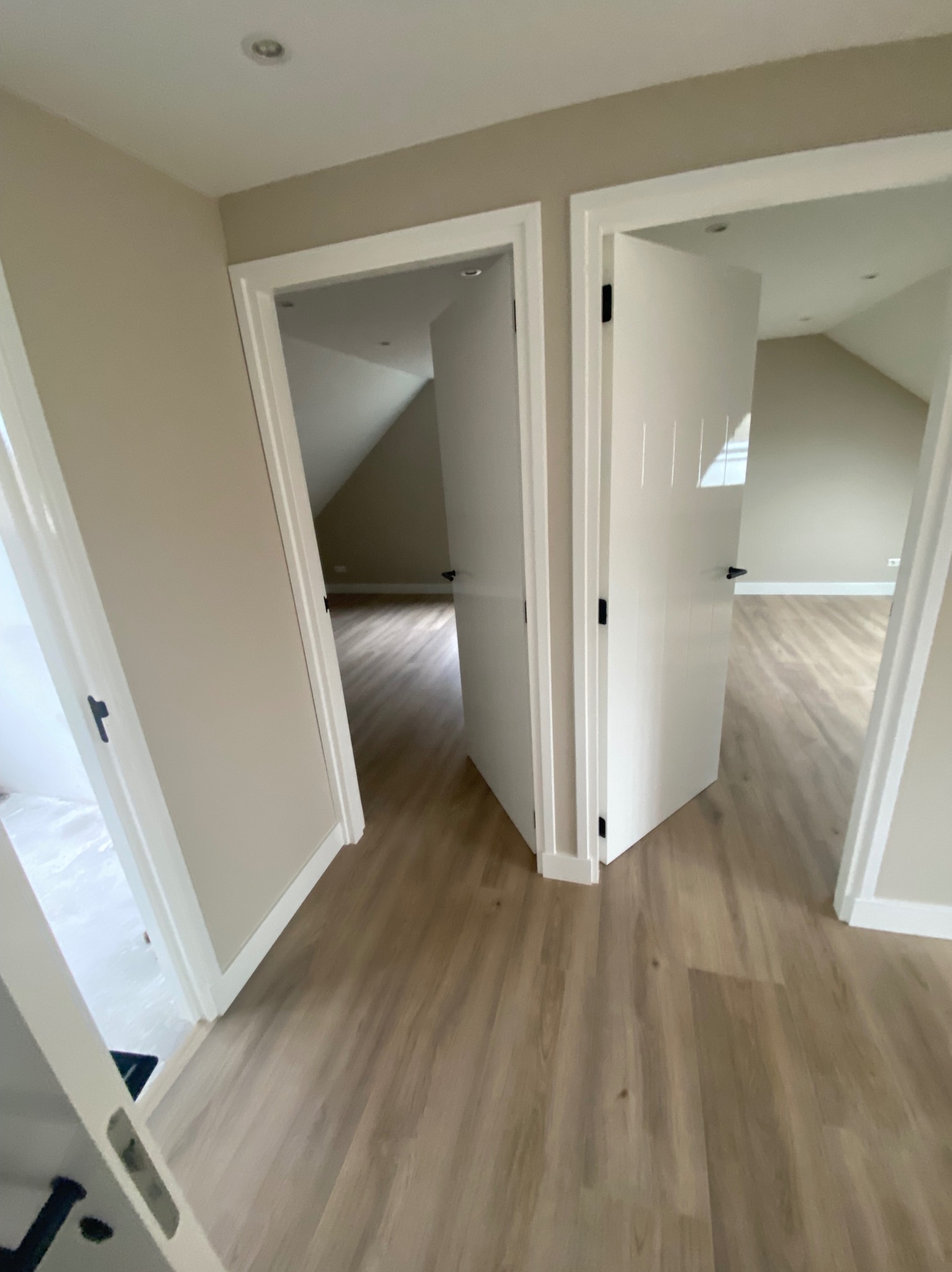 Wykończone wnętrze poddasza z drewnianą podłogą i białymi drzwiami z czarnymi klamkami, widoczne przez otwarte przejścia. Ściany w odcieniach beżu, oświetlenie punktowe.