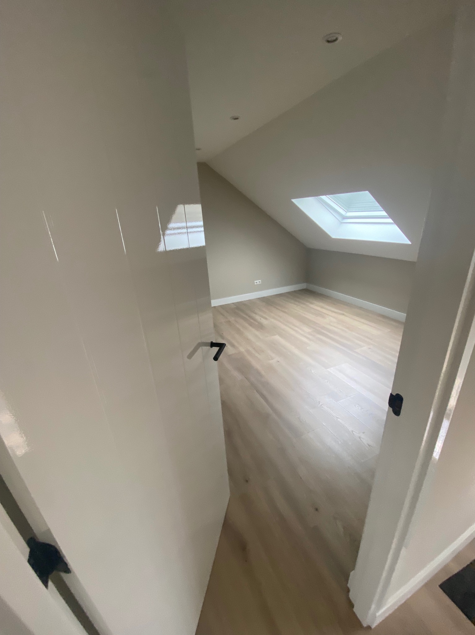 Jasne, świeżo pomalowane poddasze z oknem dachowym, widoczne przez otwarte drzwi. Parkiet na podłodze, ściany w odcieniach beżu. Minimalistyczny styl.