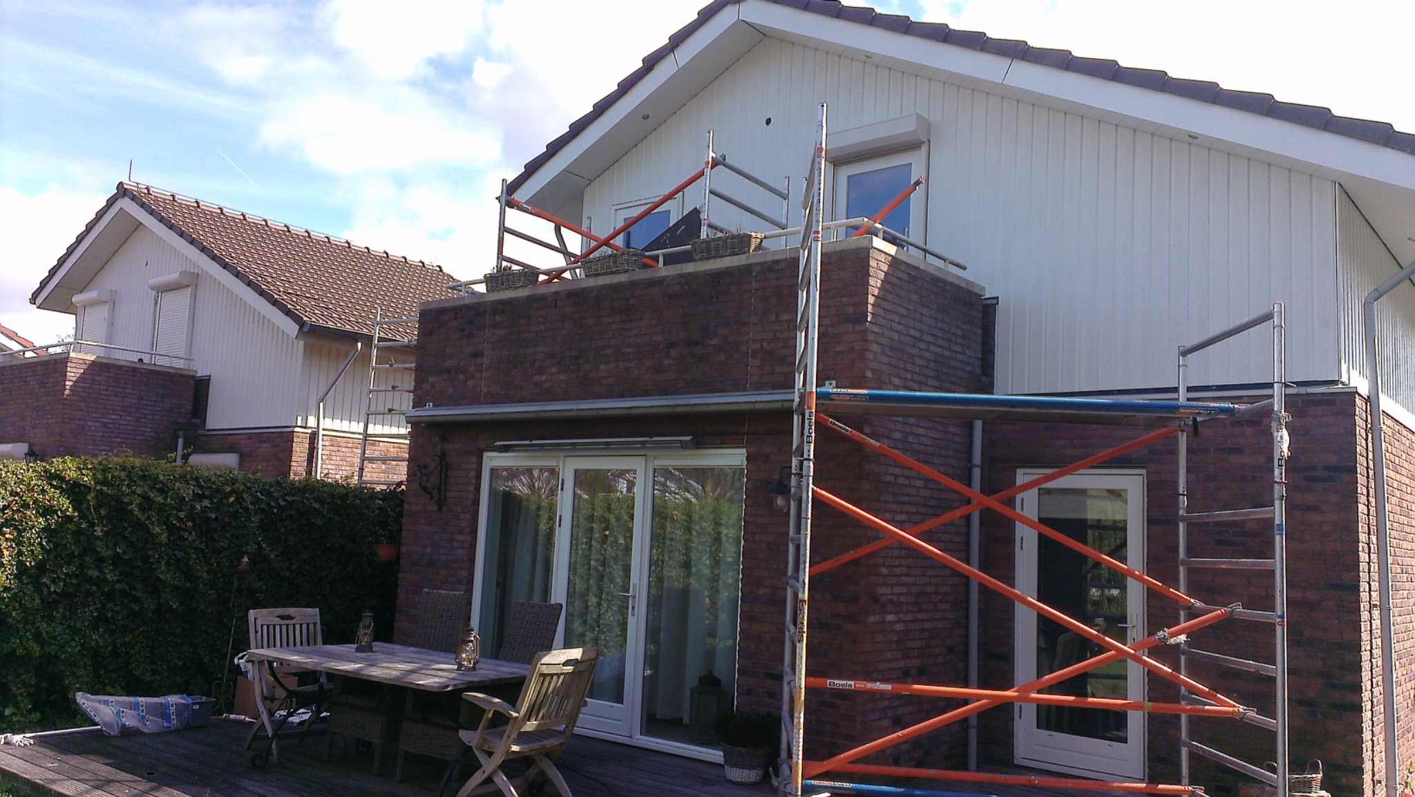 Dom z cegły i białymi panelami elewacyjnymi w trakcie prac. Widoczne rusztowania i stół ogrodowy na tarasie. Częściowo zachmurzone niebo w tle.