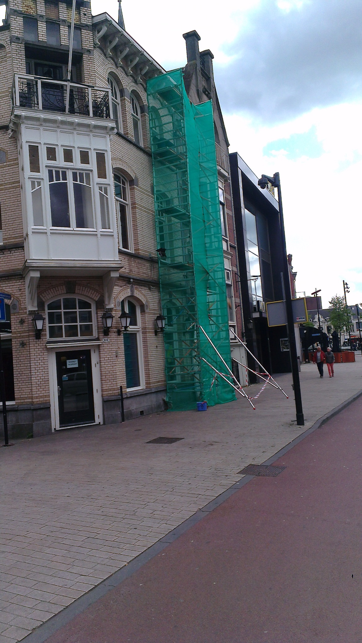 Kamienica w trakcie remontu, osłonięta zieloną siatką na rusztowaniach. Widoczny fragment chodnika i ulicy. Dwie osoby przechodzą obok.