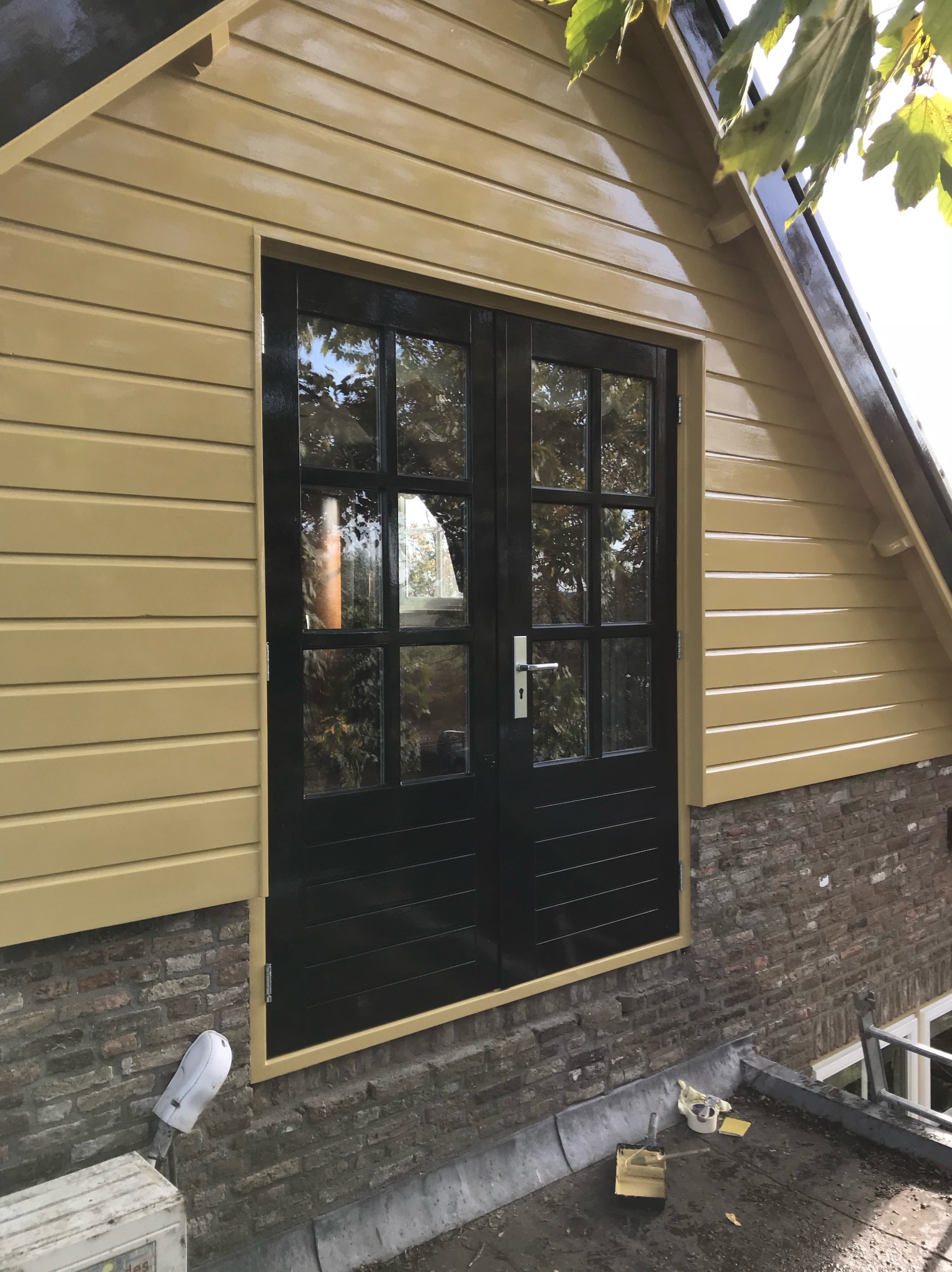 Odnowiona elewacja budynku z drewnianą okładziną w odcieniach beżu i czarnymi dwuskrzydłowymi drzwiami, widoczne narzędzia malarskie i ceglana podmurówka.