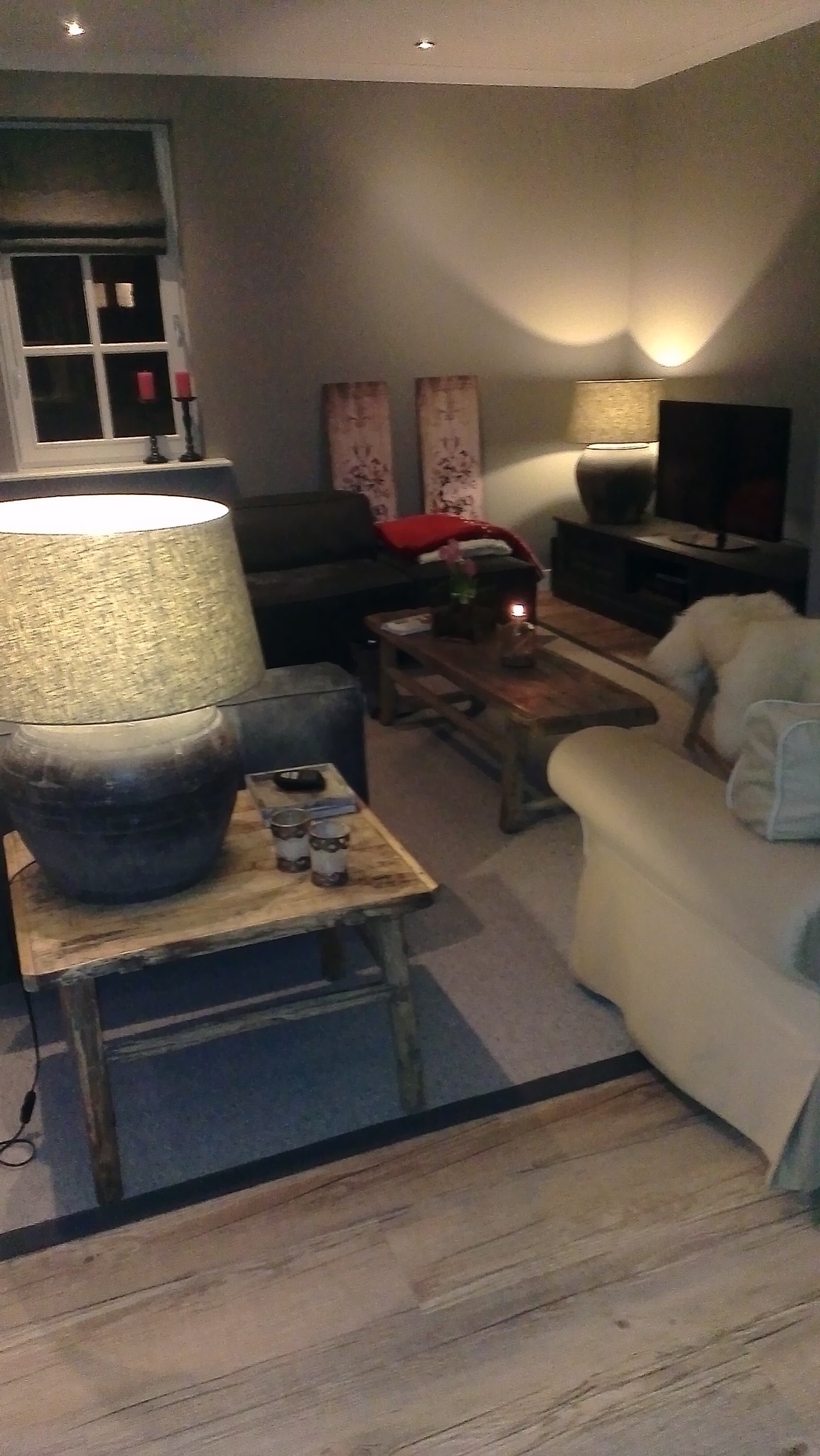 Umeblowane wnętrze salonu w neutralnych barwach. Widoczne sofy, stoliki kawowe i lampy. Ściany pomalowane na szaro, podłoga drewniana. Przytulna aranżacja.