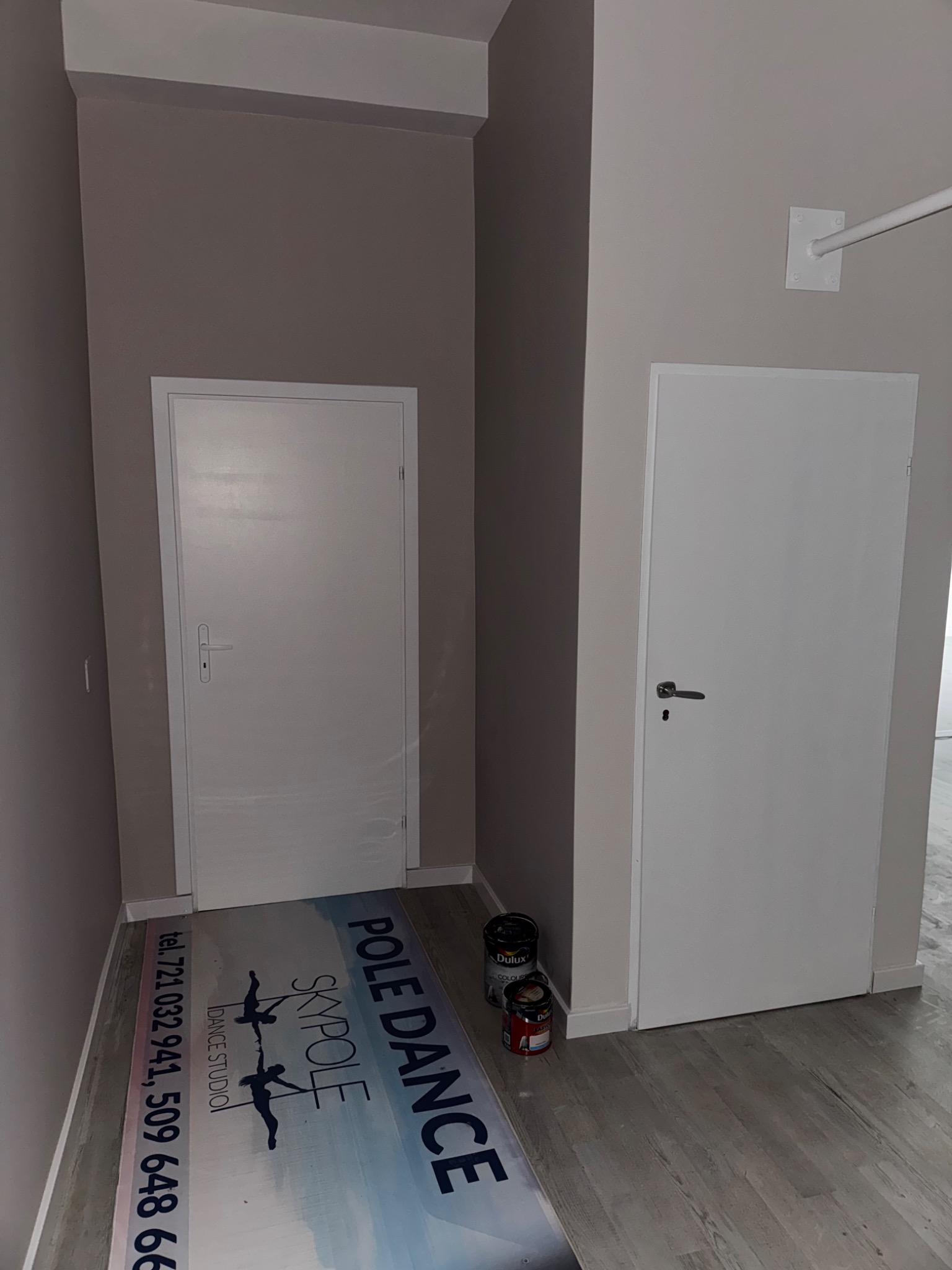 Świeżo pomalowane wnętrze z dwoma białymi drzwiami i puszkami farby Dulux. Na podłodze mata z logo studia tańca na rurze.