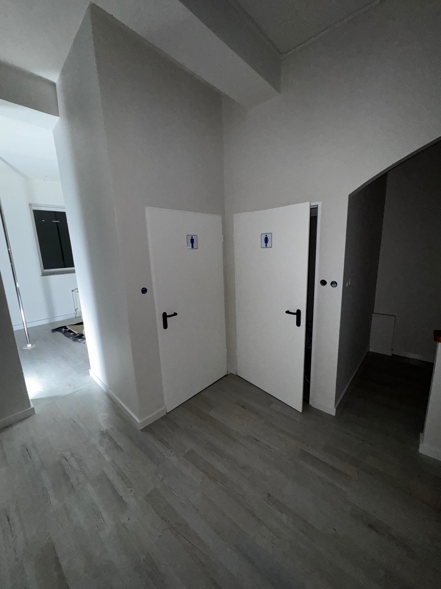Wnętrze po malowaniu: białe ściany, szara podłoga, dwie otwarte toalety z czarnymi klamkami, widoczny fragment okna i słupka do tańca.
