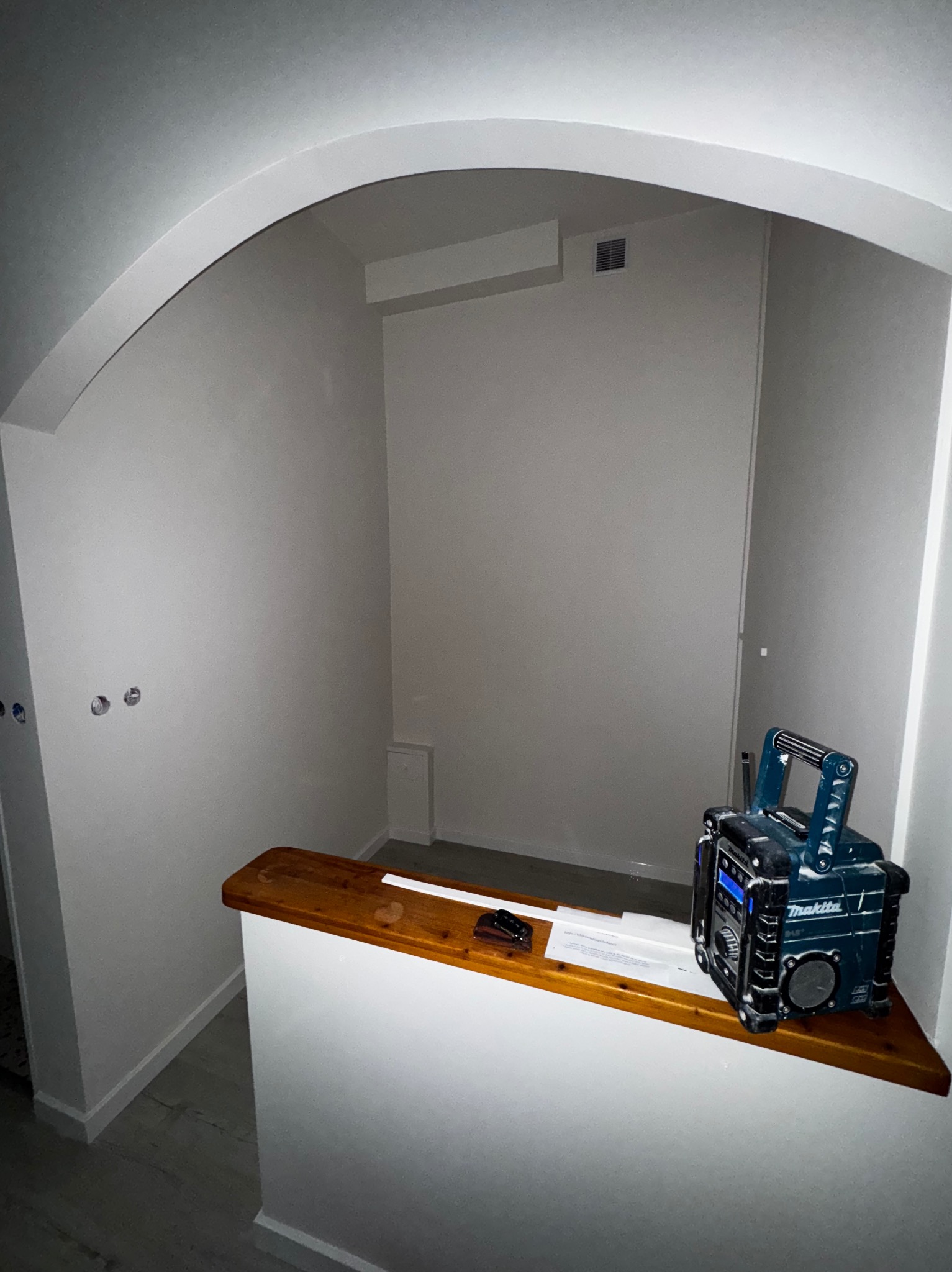 Wnęka po malowaniu na biało z radiem Makita na drewnianym blacie. Widoczny fragment instalacji elektrycznej i wentylacji. Świeżo pomalowane ściany z łukowym przejściem.