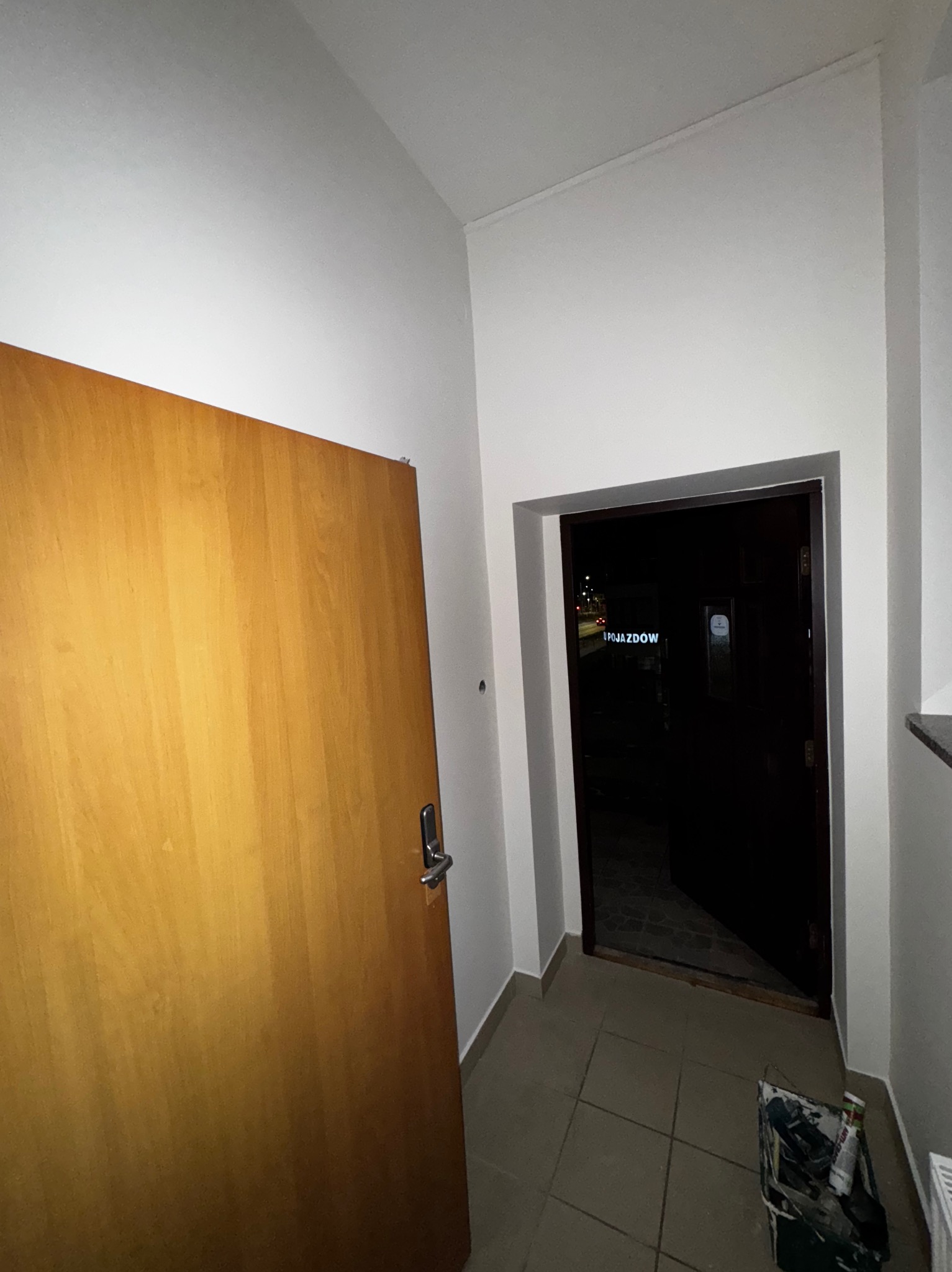 Wnętrze klatki schodowej po odświeżeniu: białe ściany, drewniane drzwi wejściowe i widok na zewnątrz przez otwarte drzwi. Na podłodze płytki i wiadro z narzędziami.