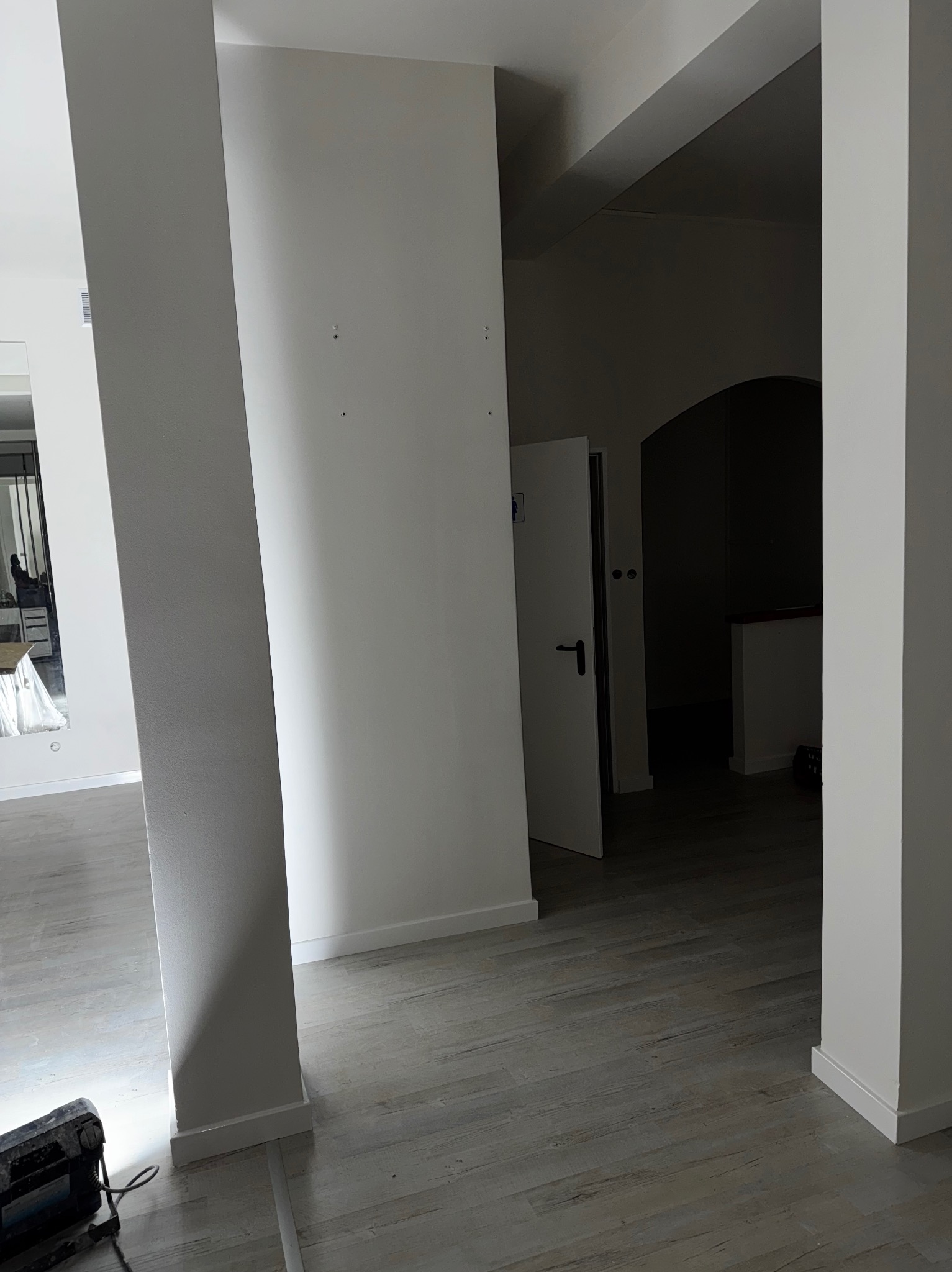Białe, świeżo pomalowane ściany i kolumny w jasnym wnętrzu. Na podłodze panele imitujące drewno. W głębi widoczne drzwi. W rogu leży lampa budowlana.