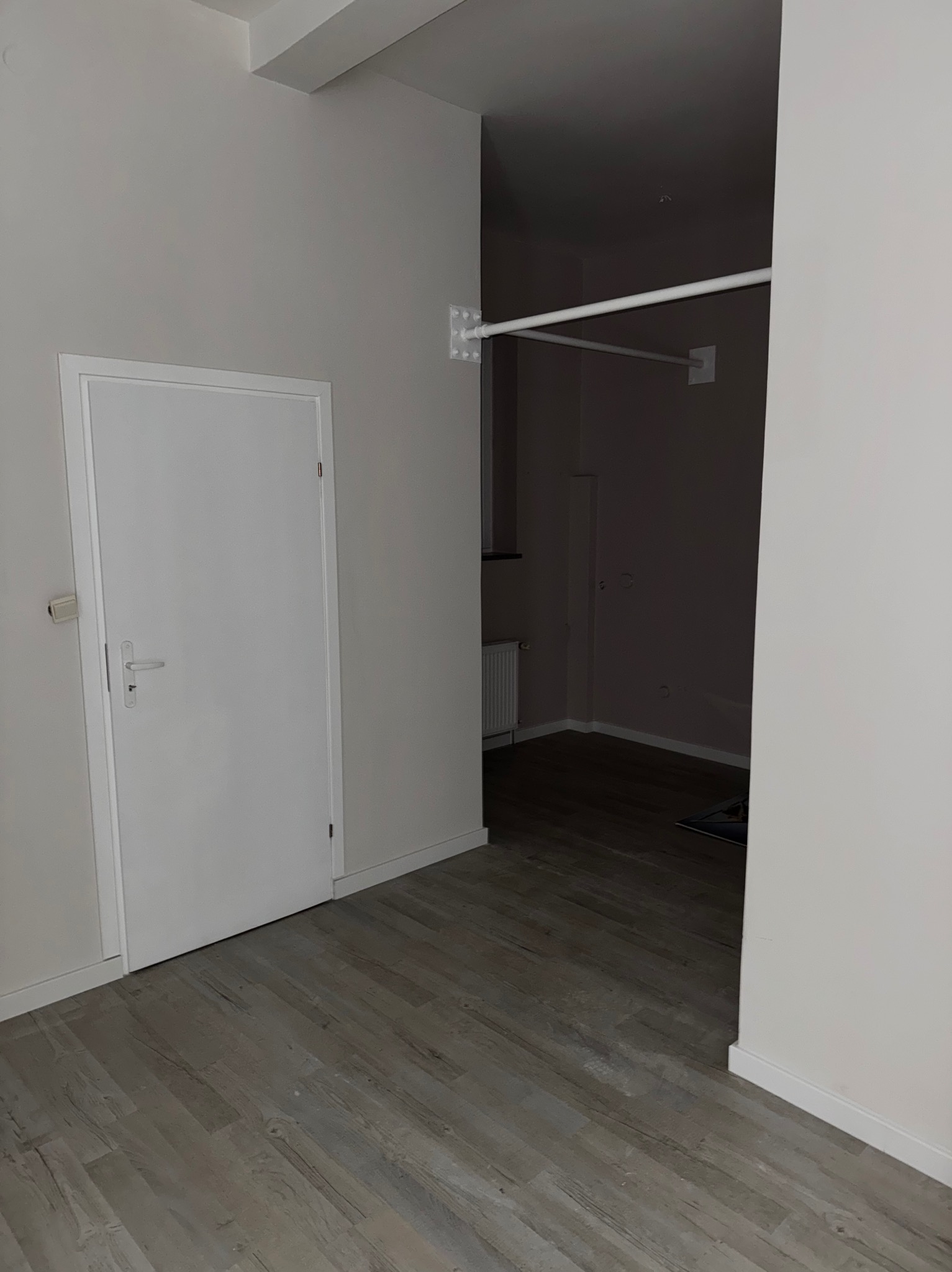 Jasne wnętrze po odświeżeniu: białe ściany, drzwi i listwy przypodłogowe, szara podłoga imitująca drewno. W tle widoczny grzejnik i rurki pod sufitem.