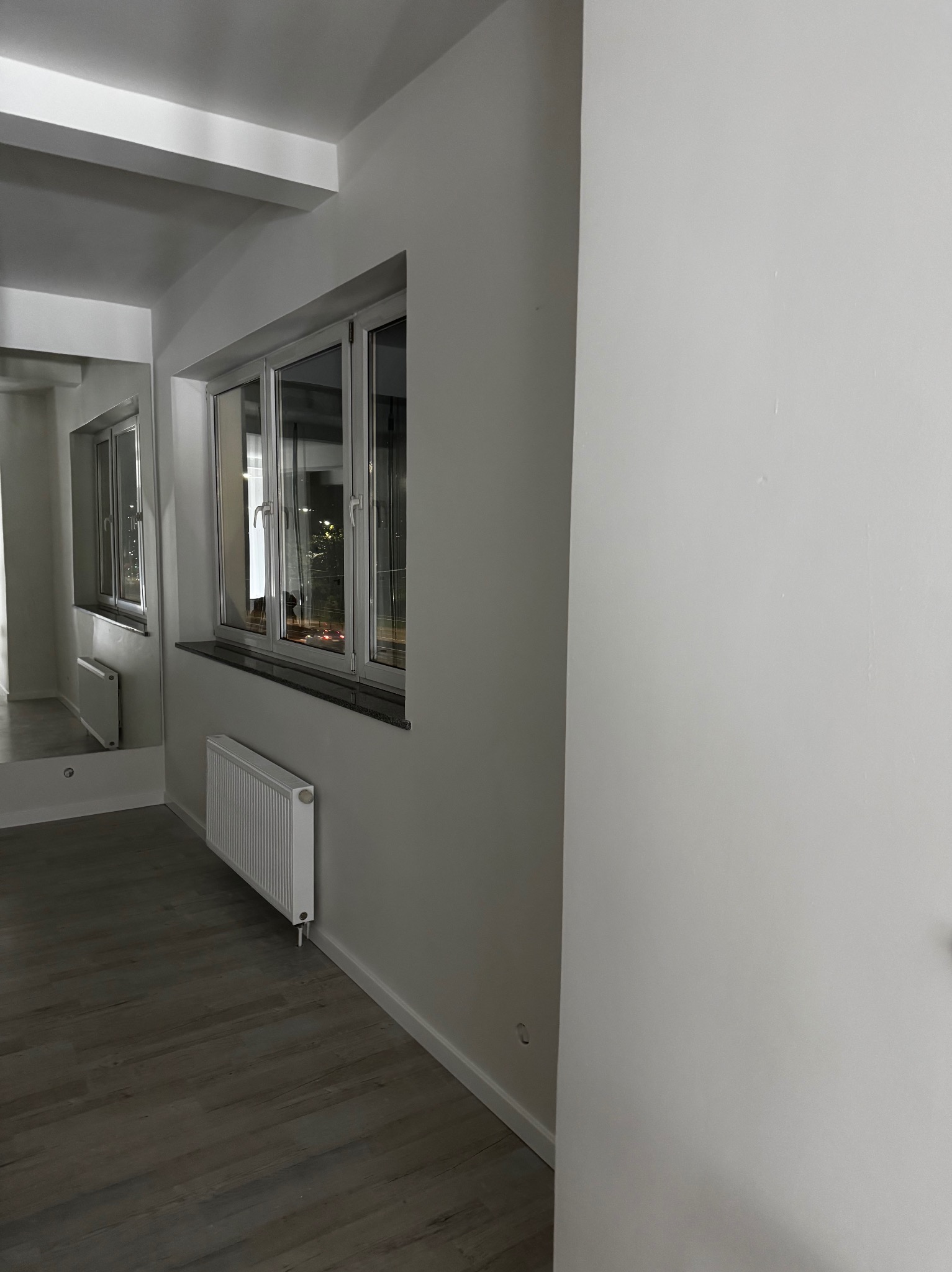 Wnętrze po odświeżeniu: gładkie, białe ściany, okno z parapetem, grzejnik. Podłoga z paneli. Widok na sąsiednie budynki odbija się w oknie i lustrze.
